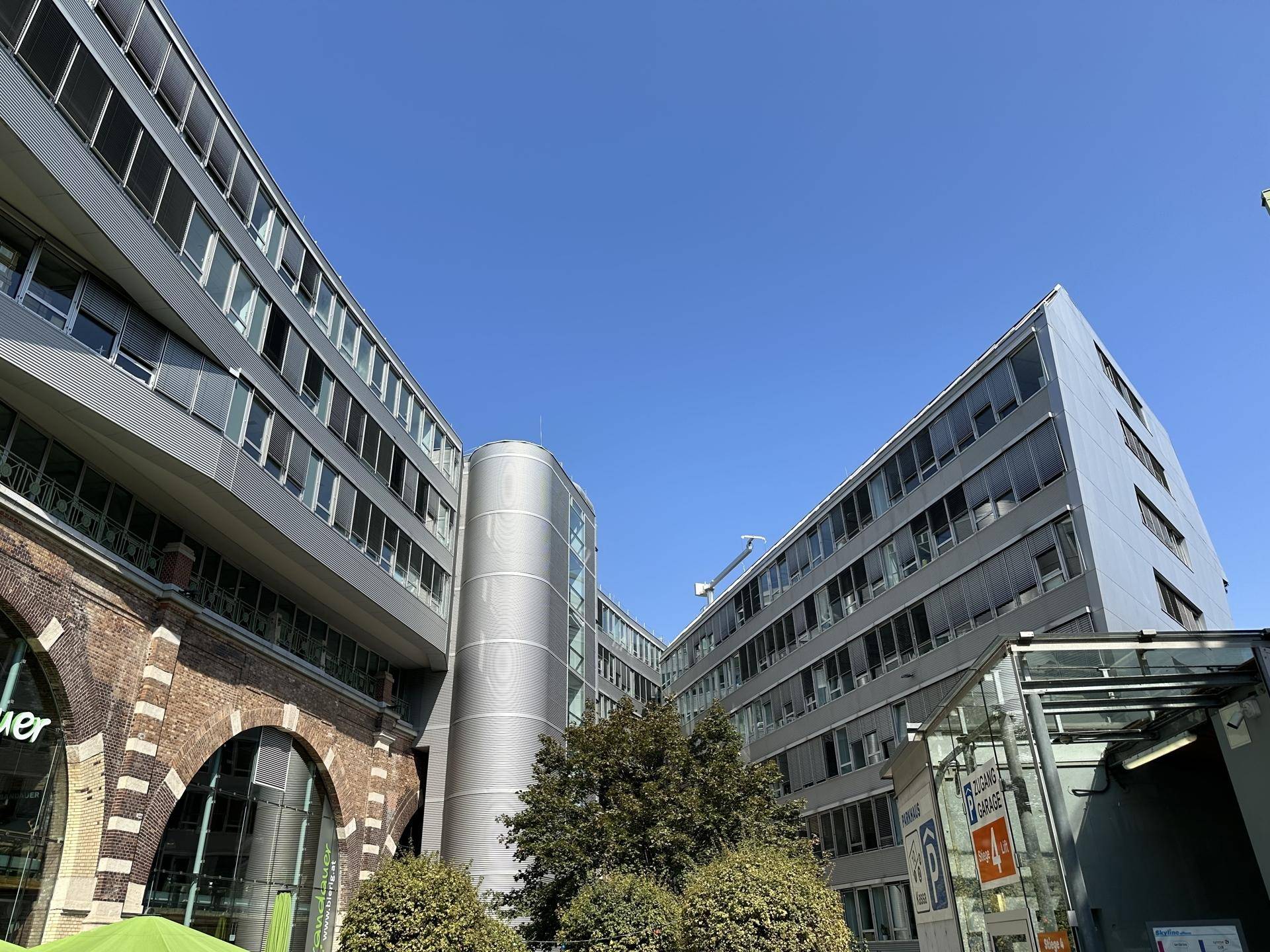 Ansicht des modernen Bürogebäudes mit Glasfassaden und historischen Backsteinbögen unter klarem Himmel.