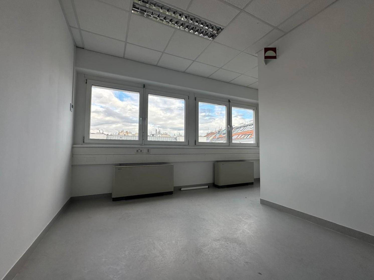 Helles Büro mit mehreren Fenstern, Heizkörpern unter der Fensterbank und grauem Bodenbelag.
