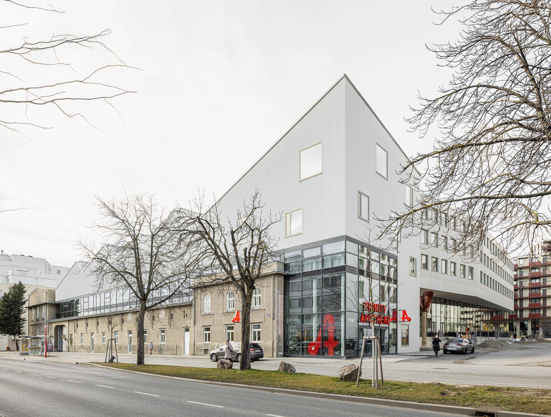 Moderne Architektur mit weißer, kantiger Fassade und großen Glasflächen an einer Straße, neben einem älteren Gebäude.