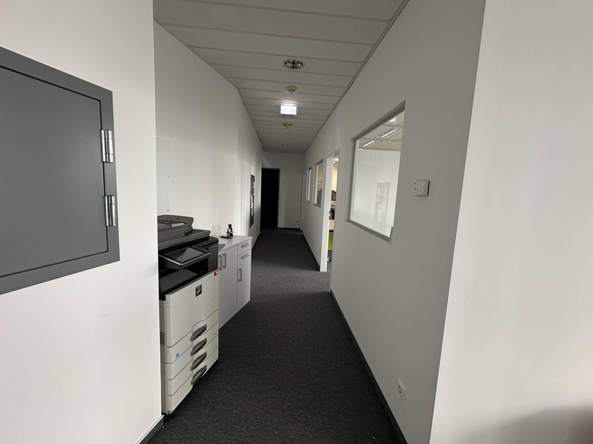 Langer, heller Flur in einem Bürogebäude mit Teppichboden, weißen Wänden und Fenstern zu angrenzenden Büros.