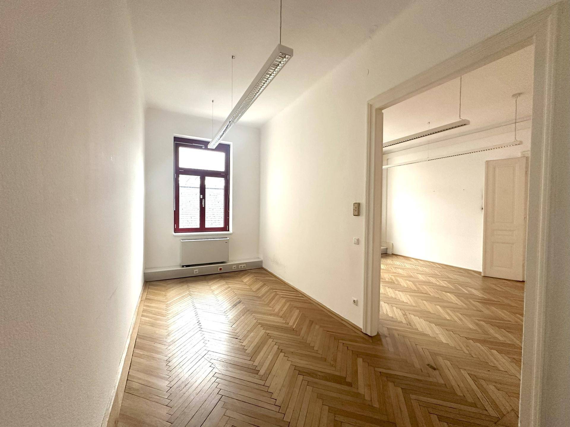 Kleinerer Büroraum mit Fischgrätparkett und einem großen Fenster, verbunden mit einem weiteren Raum.