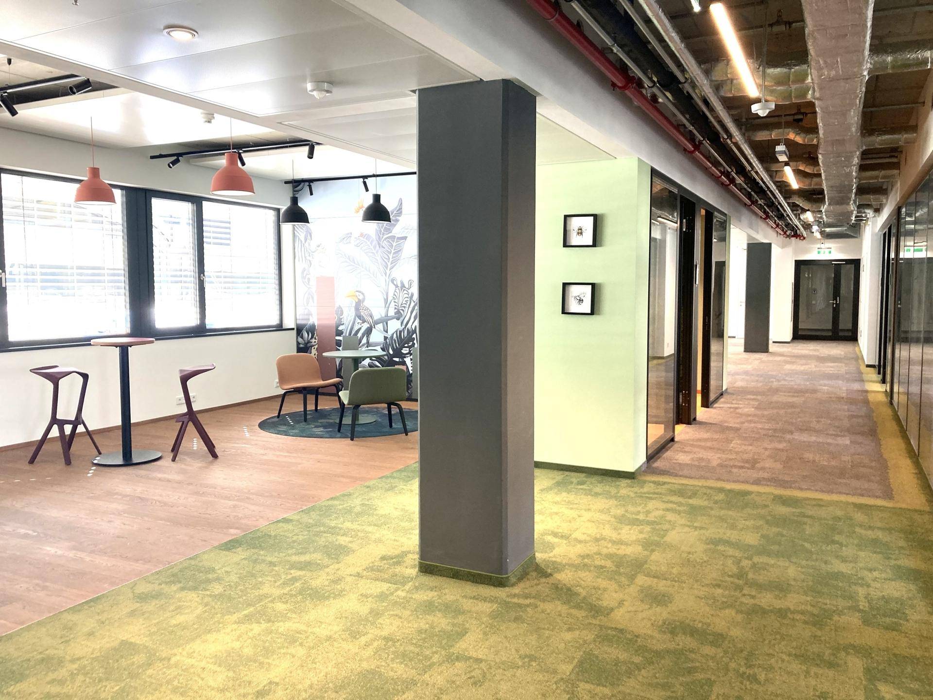 Moderner Bürobereich mit verschiedenen Sitzgelegenheiten, Teppichboden und Holzboden unter hohen Decken.