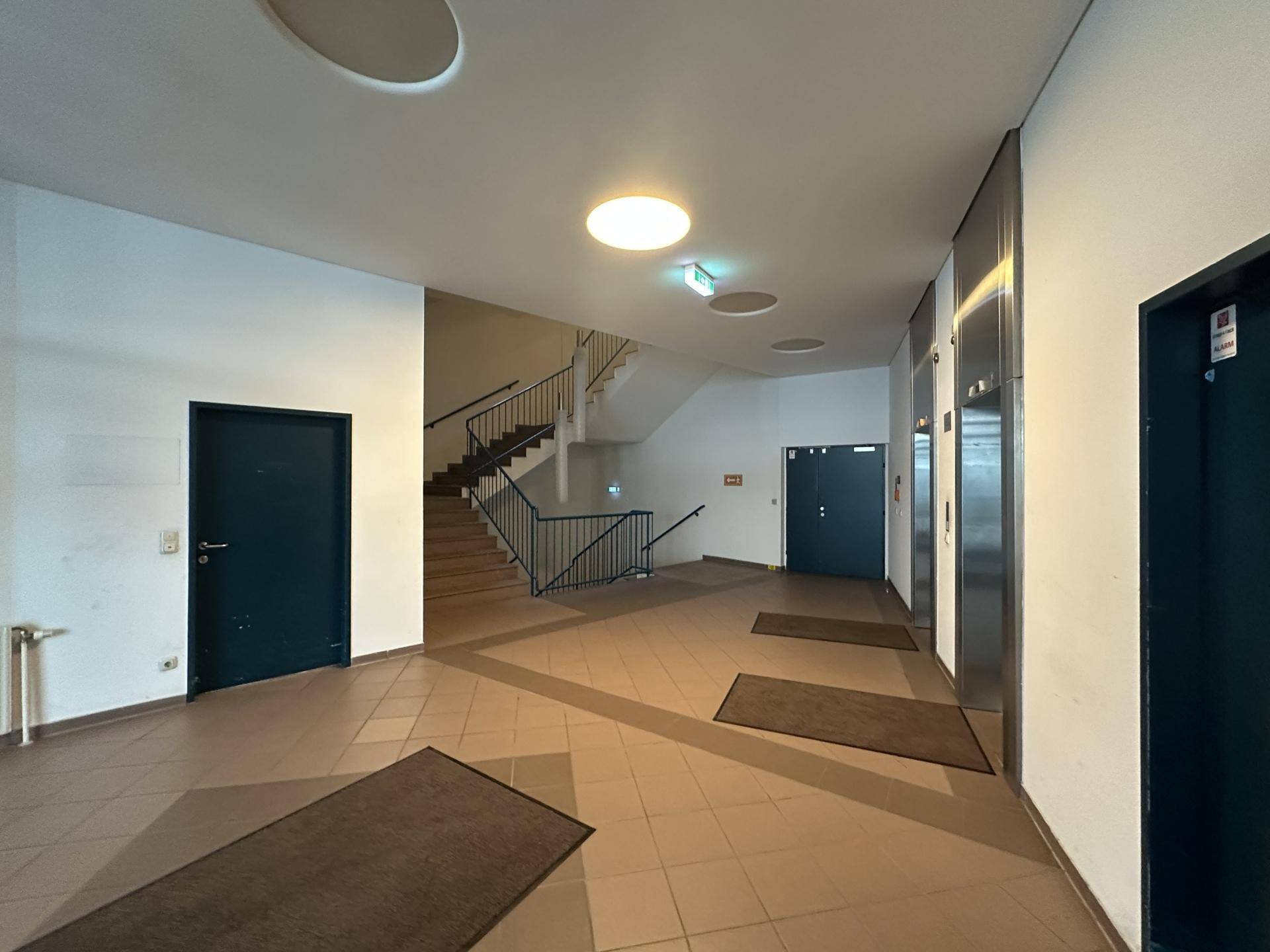 Geräumiger Eingangsbereich mit Treppenhaus, Aufzügen und braunen Fliesenböden.