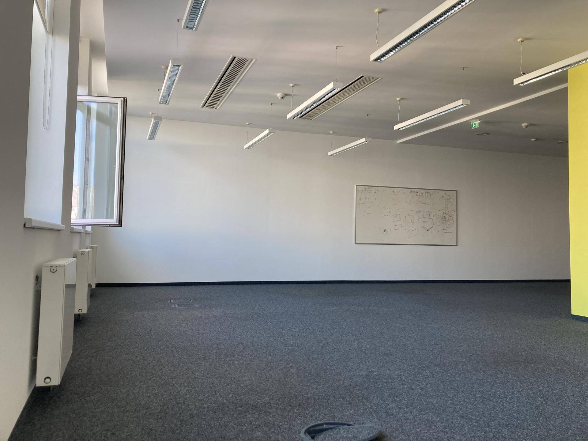 Leeres Büro mit Teppichboden, weißen Wänden, einem Whiteboard und Deckenbeleuchtung.