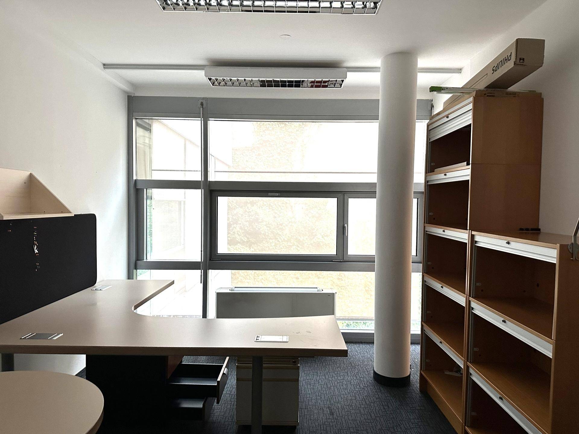 Büro mit Teppichboden, großem Fenster und funktionalen Büromöbeln, bereit zur Einrichtung.