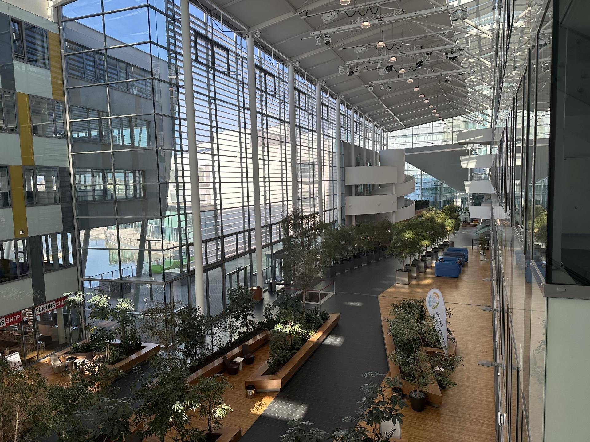 Helles, modernes Atrium mit hohen Glaswänden, vielen Pflanzen und Blick auf ein Gewässer im Außenbereich.