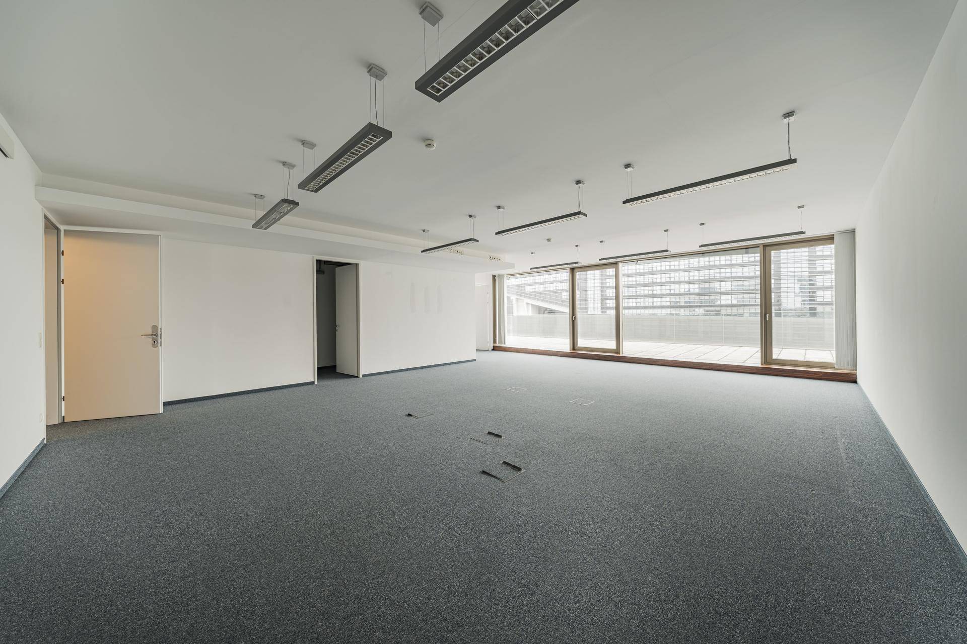 Geräumiges, leeres Büro mit Teppichboden und großen Fenstern, die auf eine Terrasse führen.
