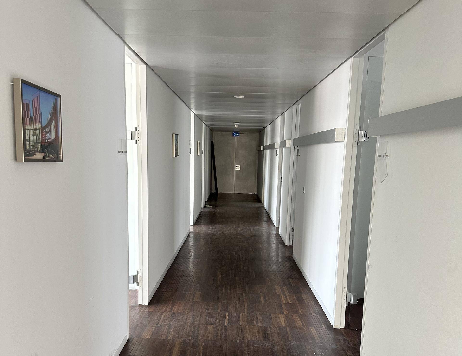 Langer, heller Flur mit weißen Wänden und einem dunklen Holzparkettboden, der zu mehreren Büros führt.