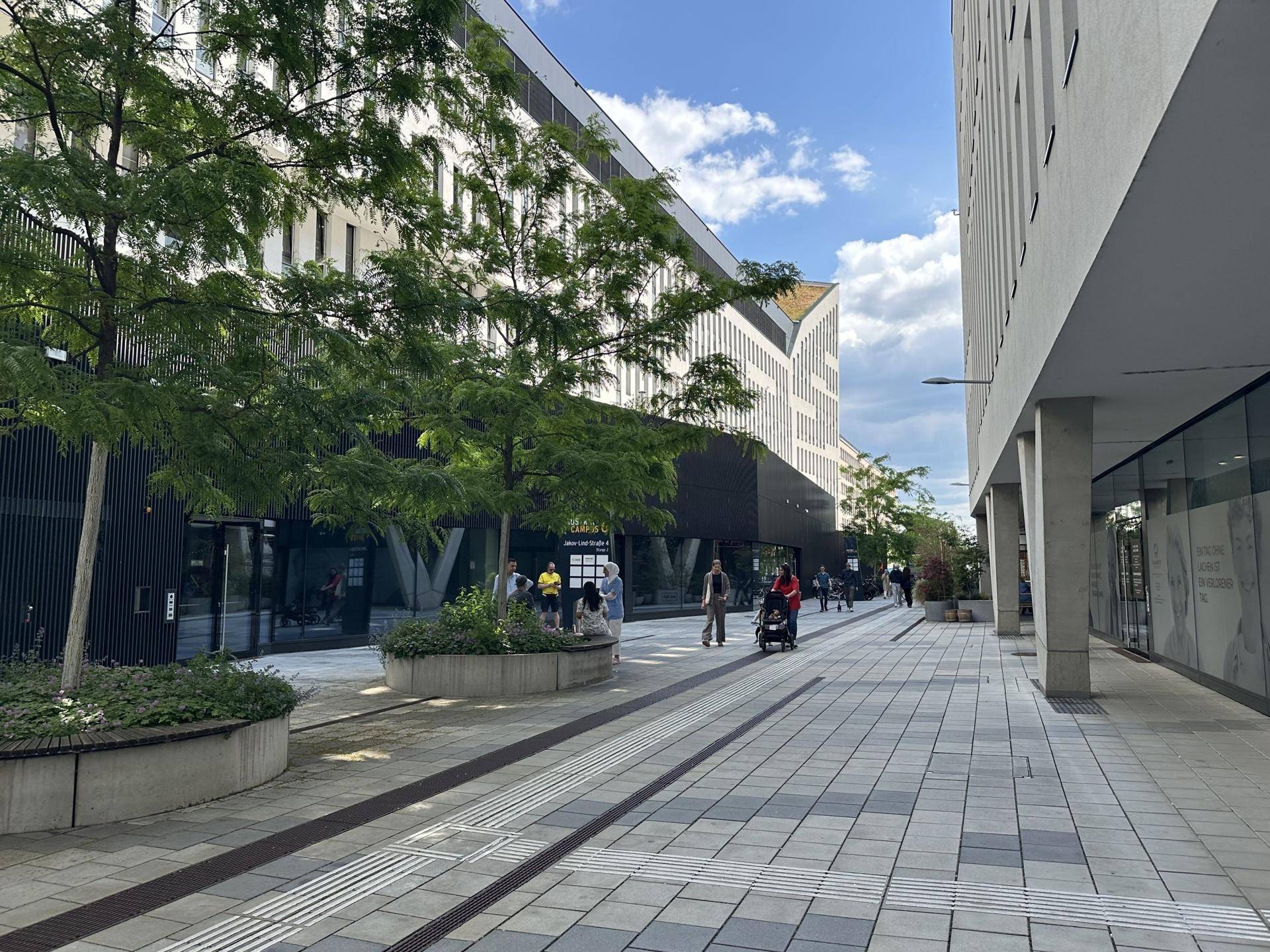 Belebte moderne Straße mit Bäumen, Sitzgelegenheiten und zeitgenössischen Gebäuden unter blauem Himmel.