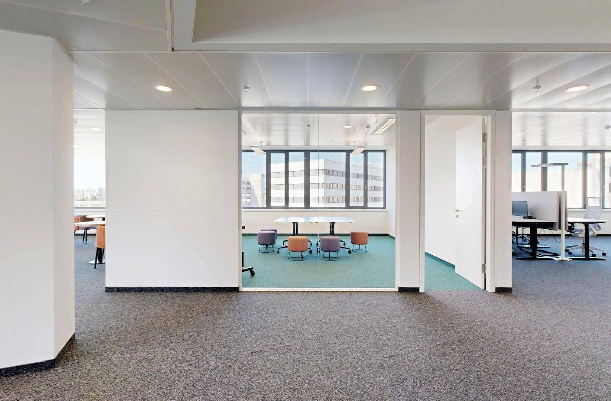 Offener Bürobereich mit Teppichboden und einem abgetrennten Besprechungsbereich mit bunten Hockern.