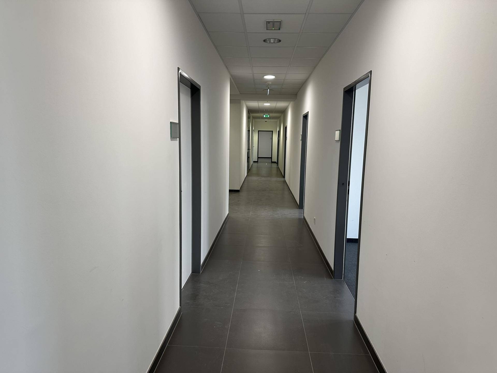 Langer, heller Flur mit grauen Fliesen, weißen Wänden und mehreren Türen zu Büroräumen.