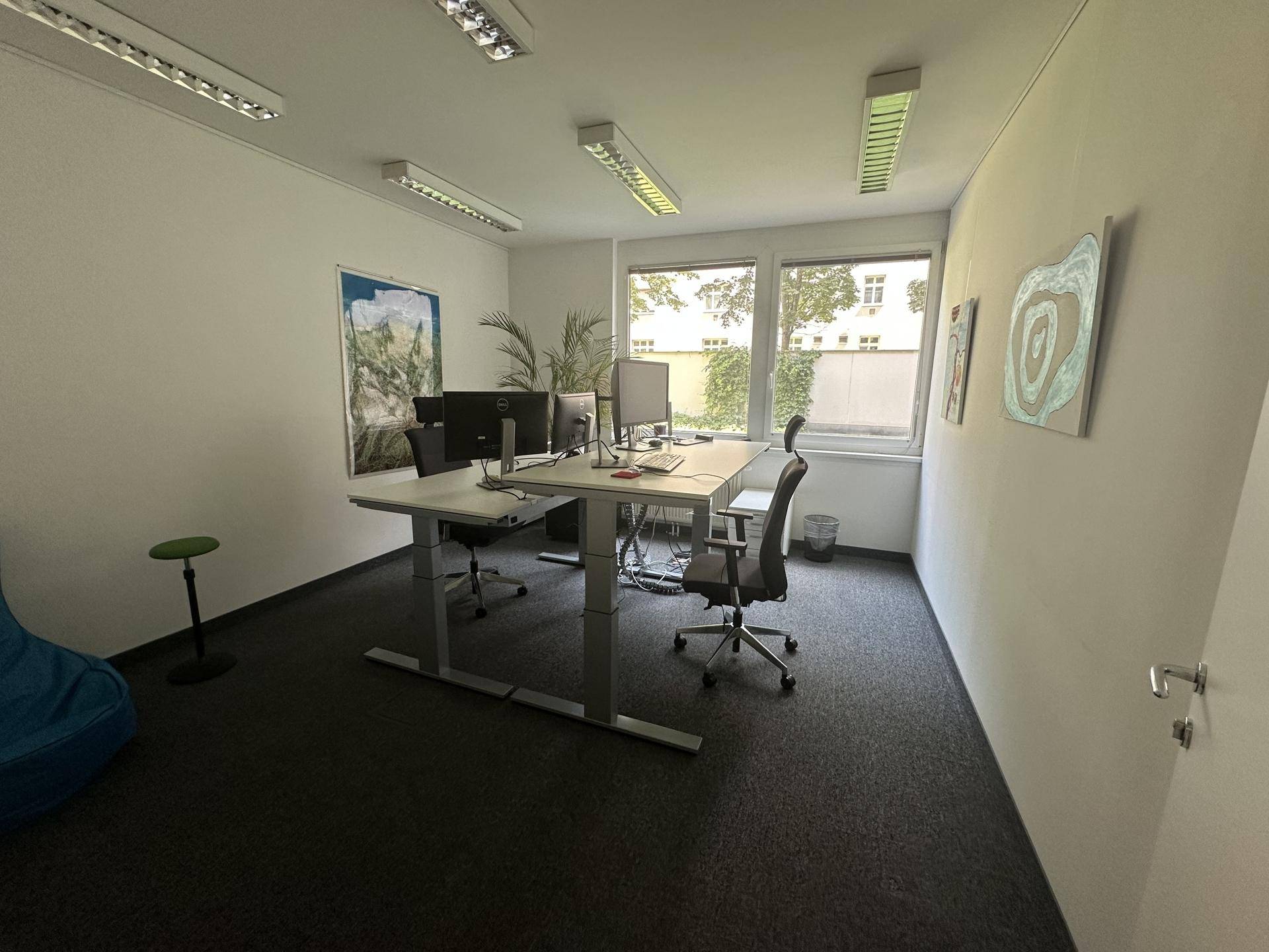Helles Büro mit zwei Schreibtischen, ergonomischen Stühlen und Teppichboden, bietet Blick ins Grüne.