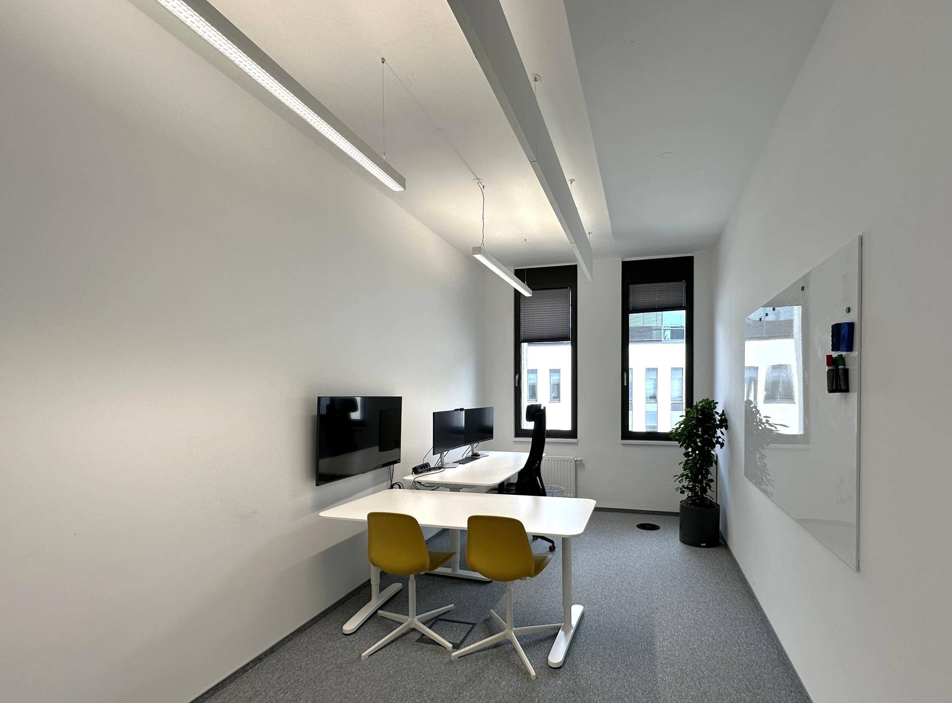 Modernes Büro mit zwei Arbeitsplätzen, gelben Stühlen, grauen Teppichböden und einer Zimmerpflanze am Fenster.