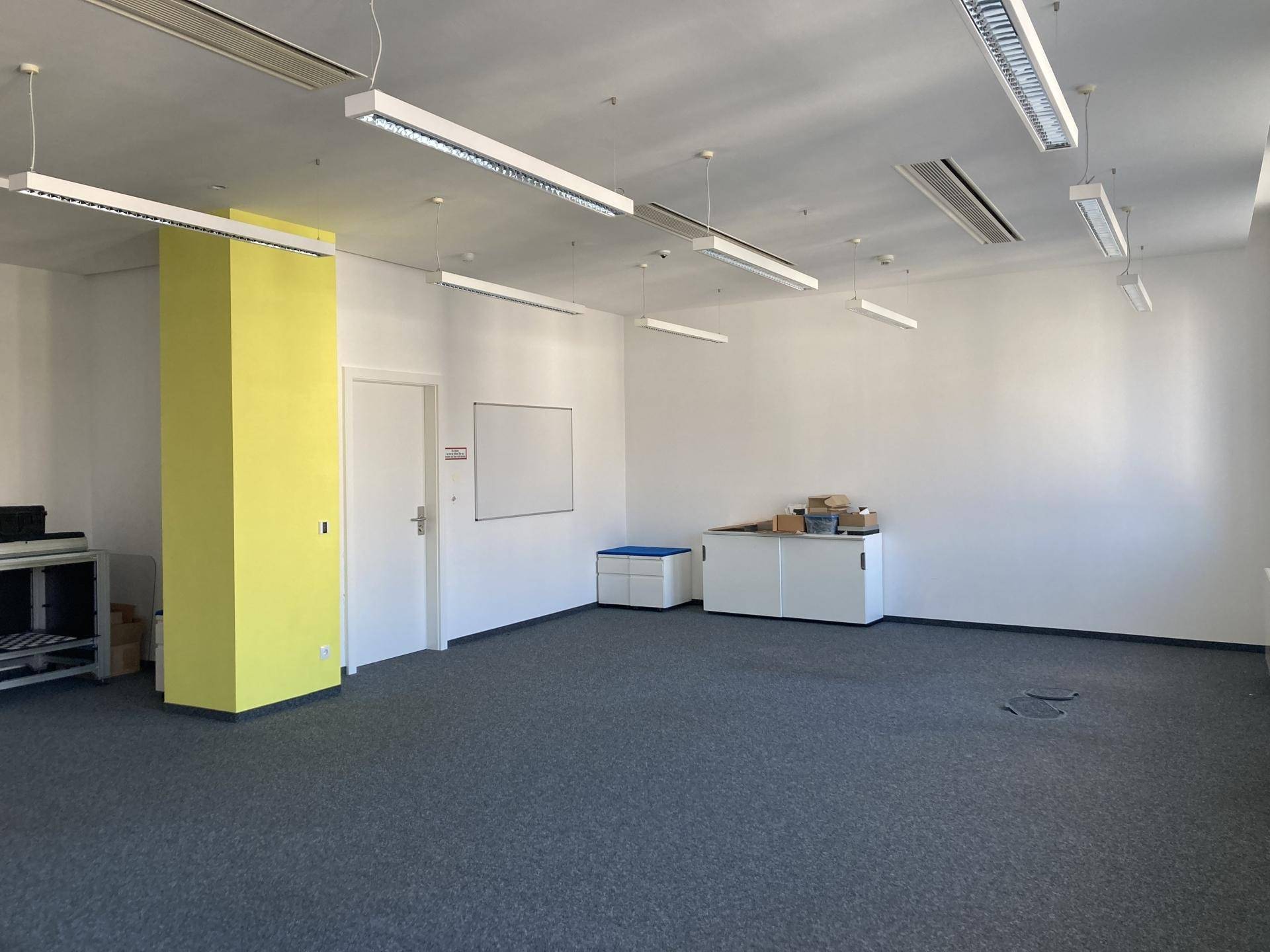 Geräumiges Büro mit Teppichboden, einem gelben Pfeiler und weißen Schränken an der Wand.