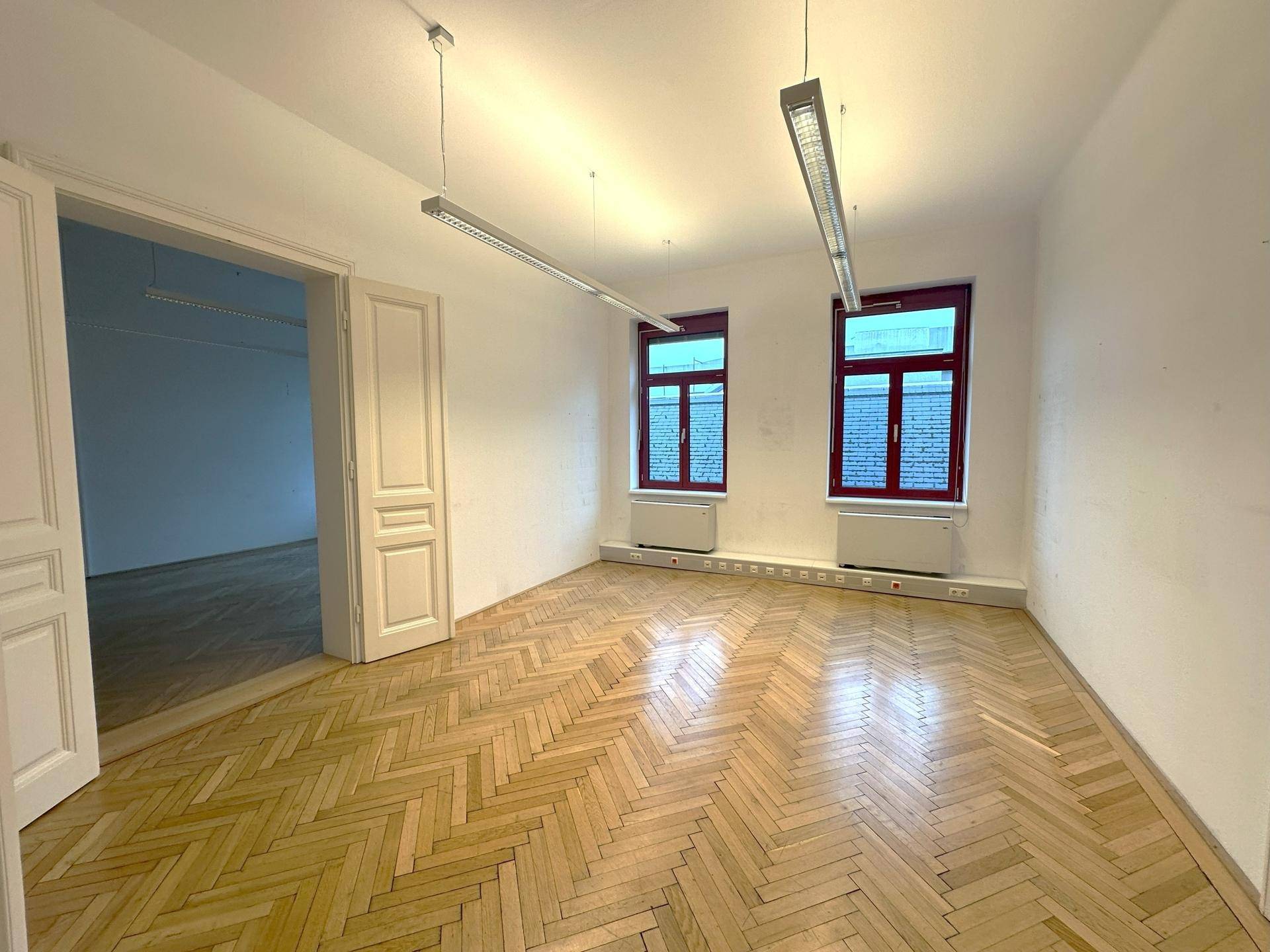 Heller Büroraum mit klassischem Fischgrätparkett und hohen Fenstern, ideal für Arbeitsbereiche.
