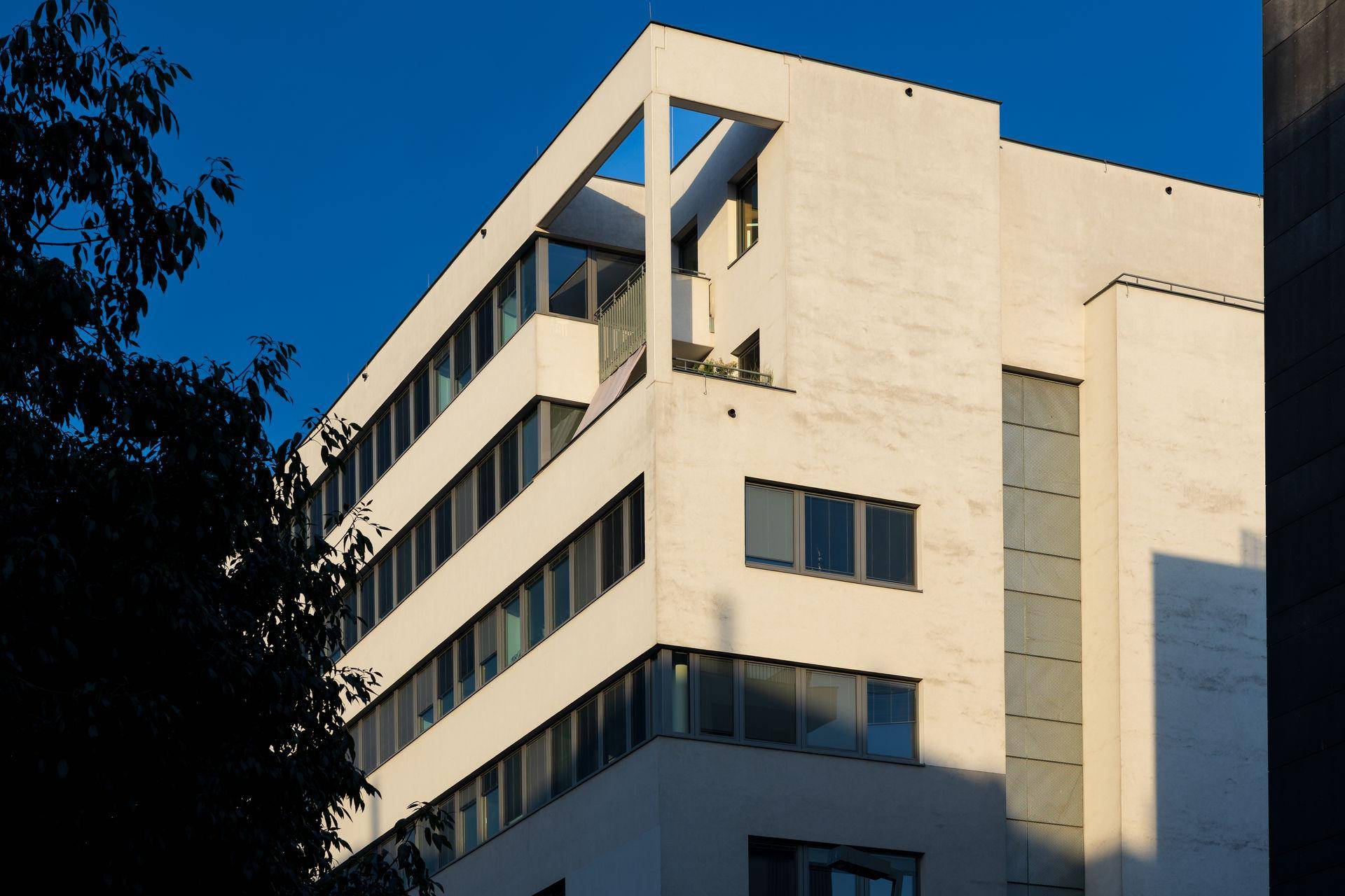 Oberer Teil eines modernen Gebäudes mit klaren Linien und großen Fenstern unter blauem Himmel bei Sonnenschein.