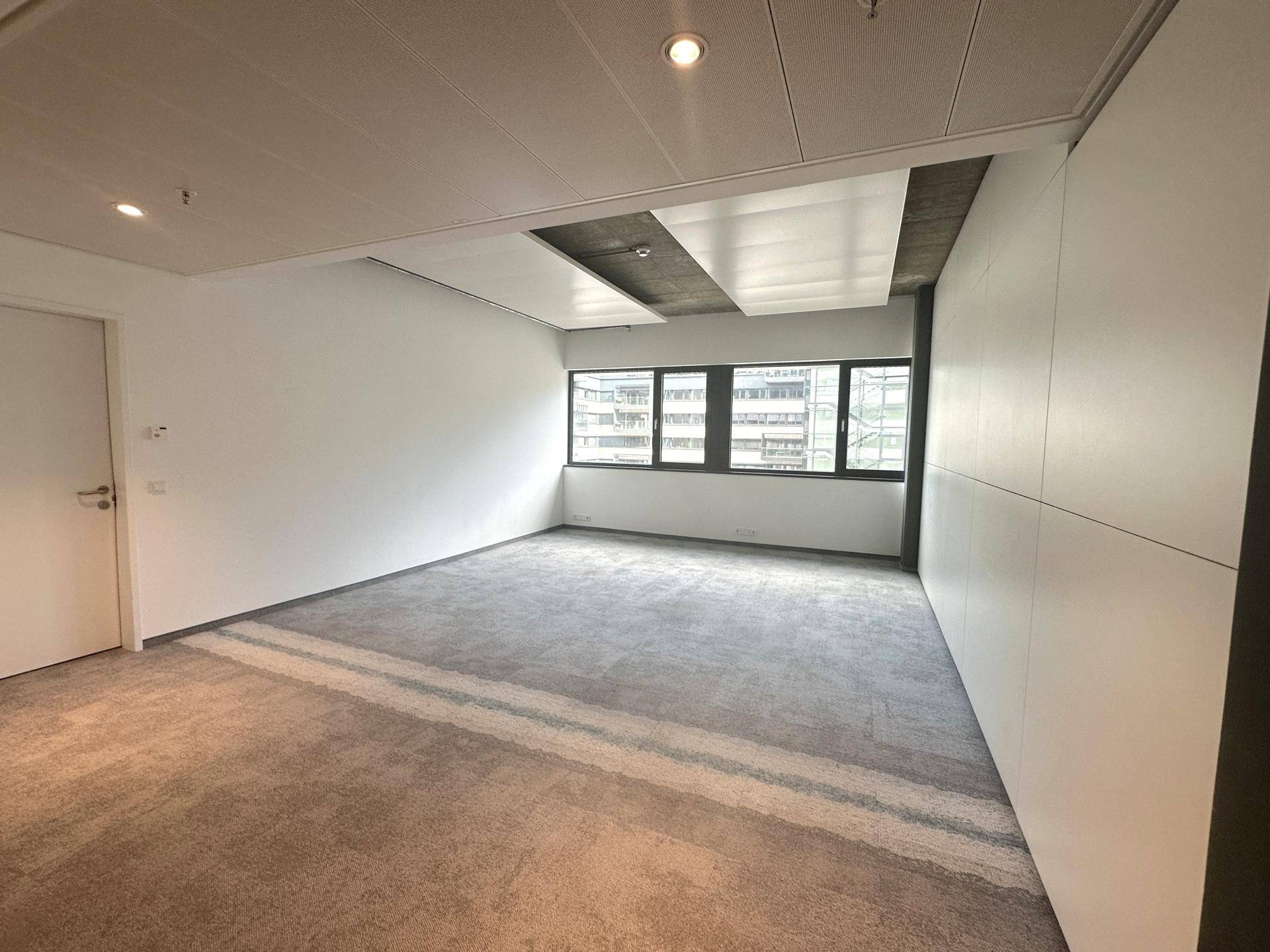 Heller Büroraum mit Teppichboden, großen Fenstern und einer modernen Deckenkonstruktion.