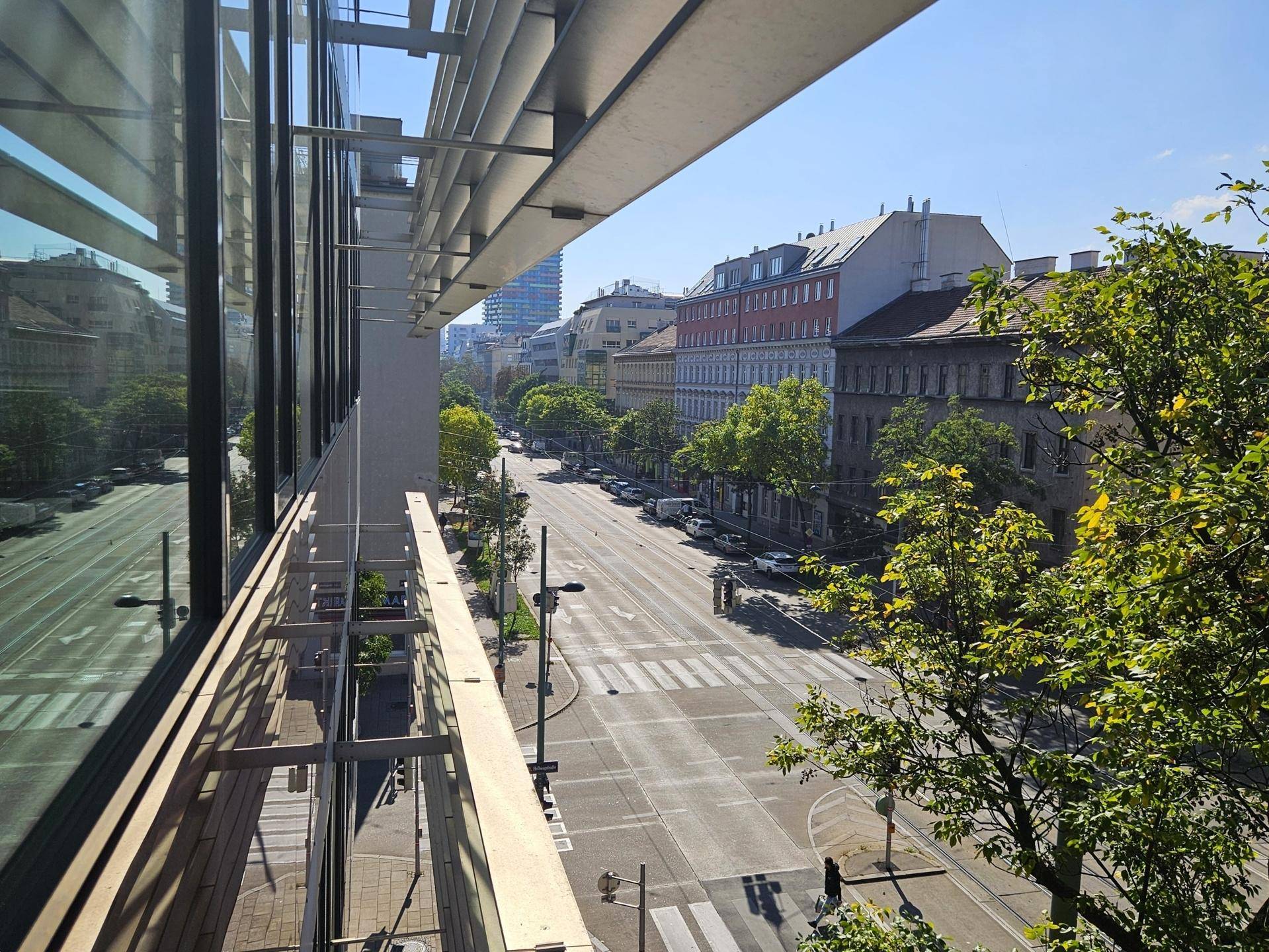 Blick von einem modernen Gebäude auf eine belebte Stadtstraße mit Bäumen und historischen Gebäuden unter blauem Himmel.