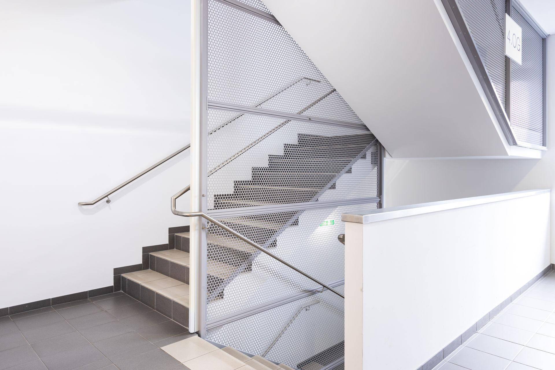 Modernes Treppenhaus mit Metallgeländer und grauen Fliesenstufen in einem hellen Gebäude mit weißen Wänden.