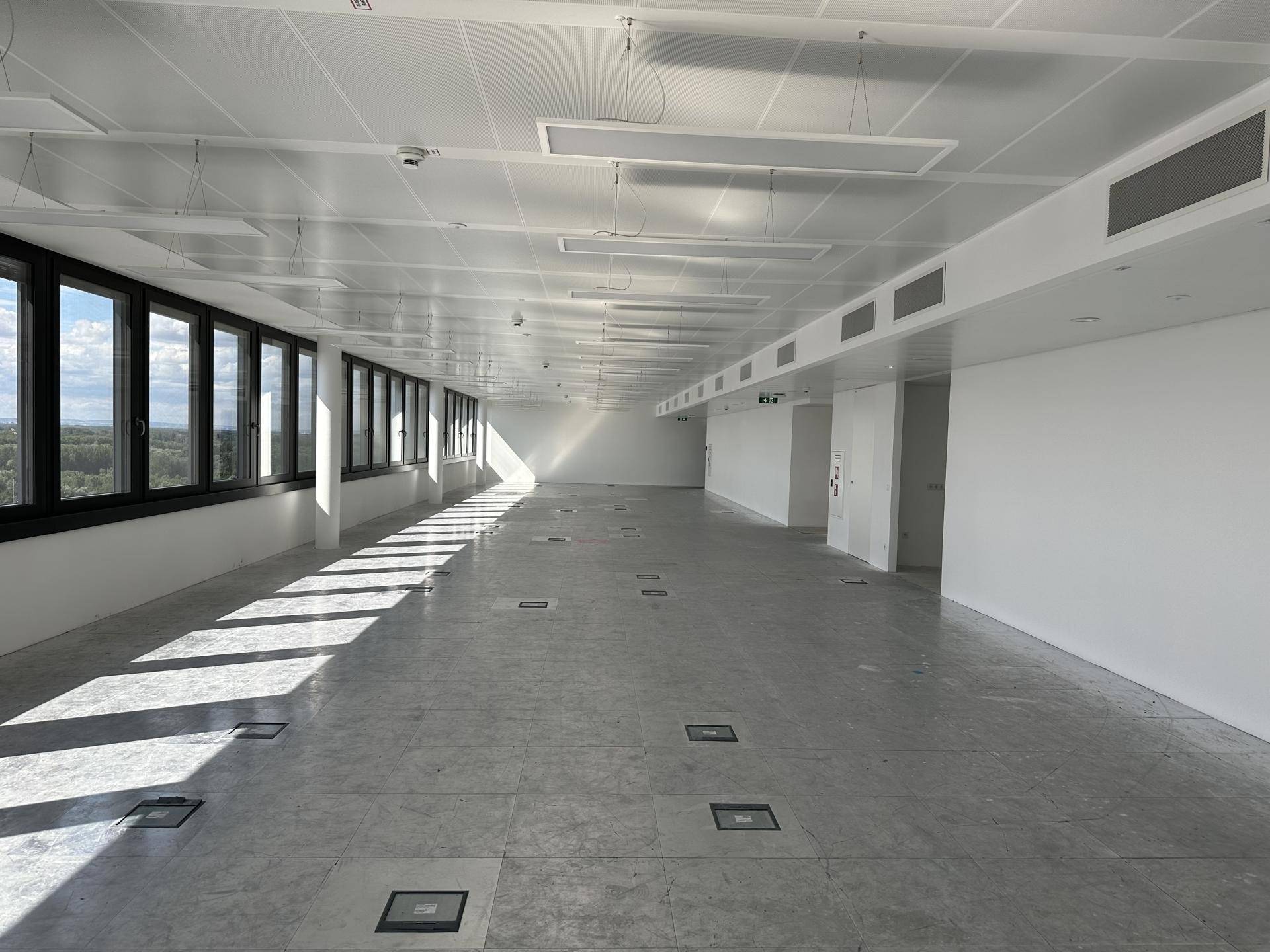 Großer, leerer Büroraum mit vielen Fenstern, hellen Deckenplatten und Bodentanks für Anschlüsse.