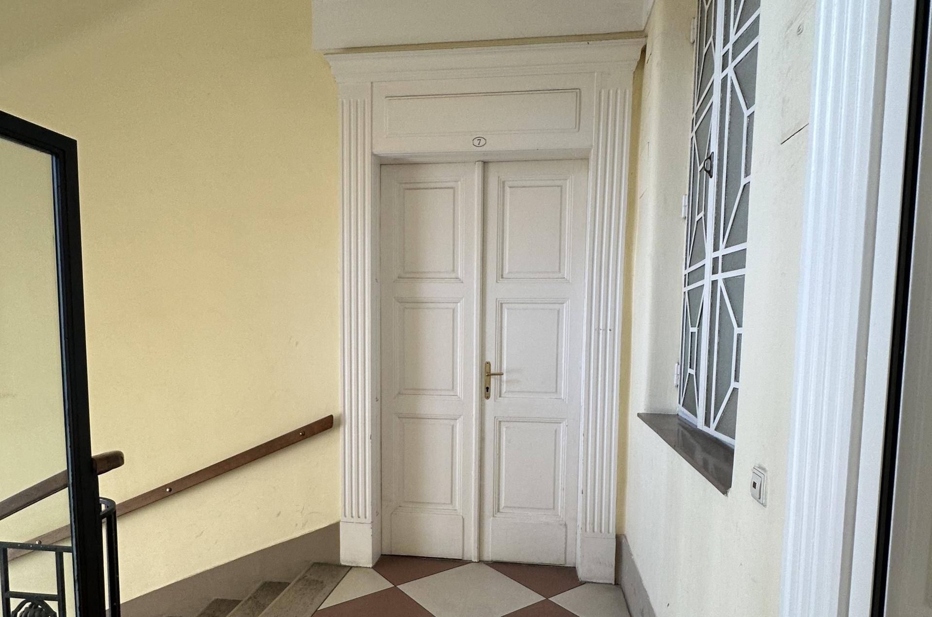 Treppenhausabsatz mit einer verzierten weißen Doppeltür und einem Fenster mit Gittermuster.