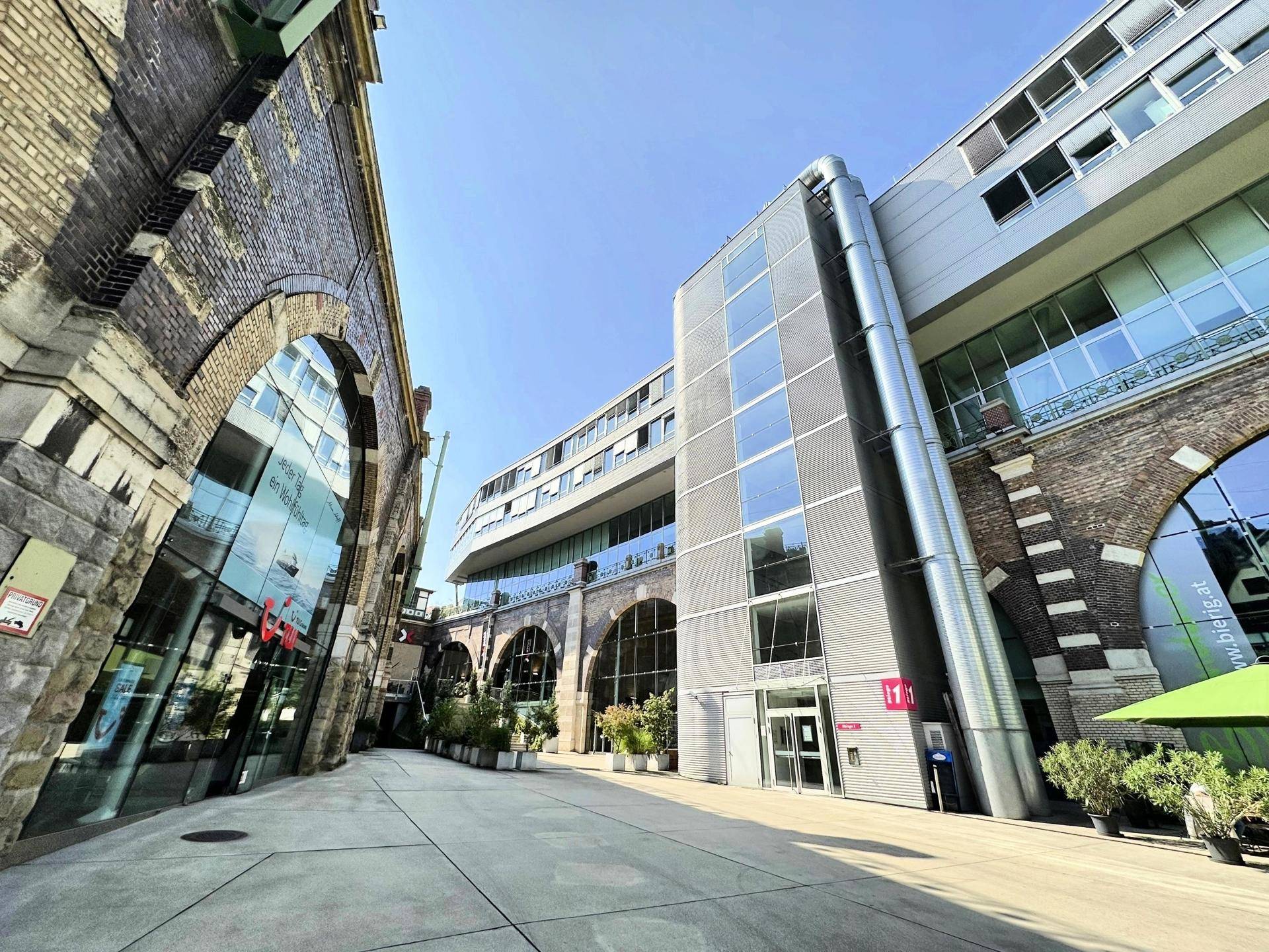 Eingangsbereich eines Gebäudekomplexes, der moderne Architektur mit historischen Backsteinbögen und Glasfassaden verbindet.