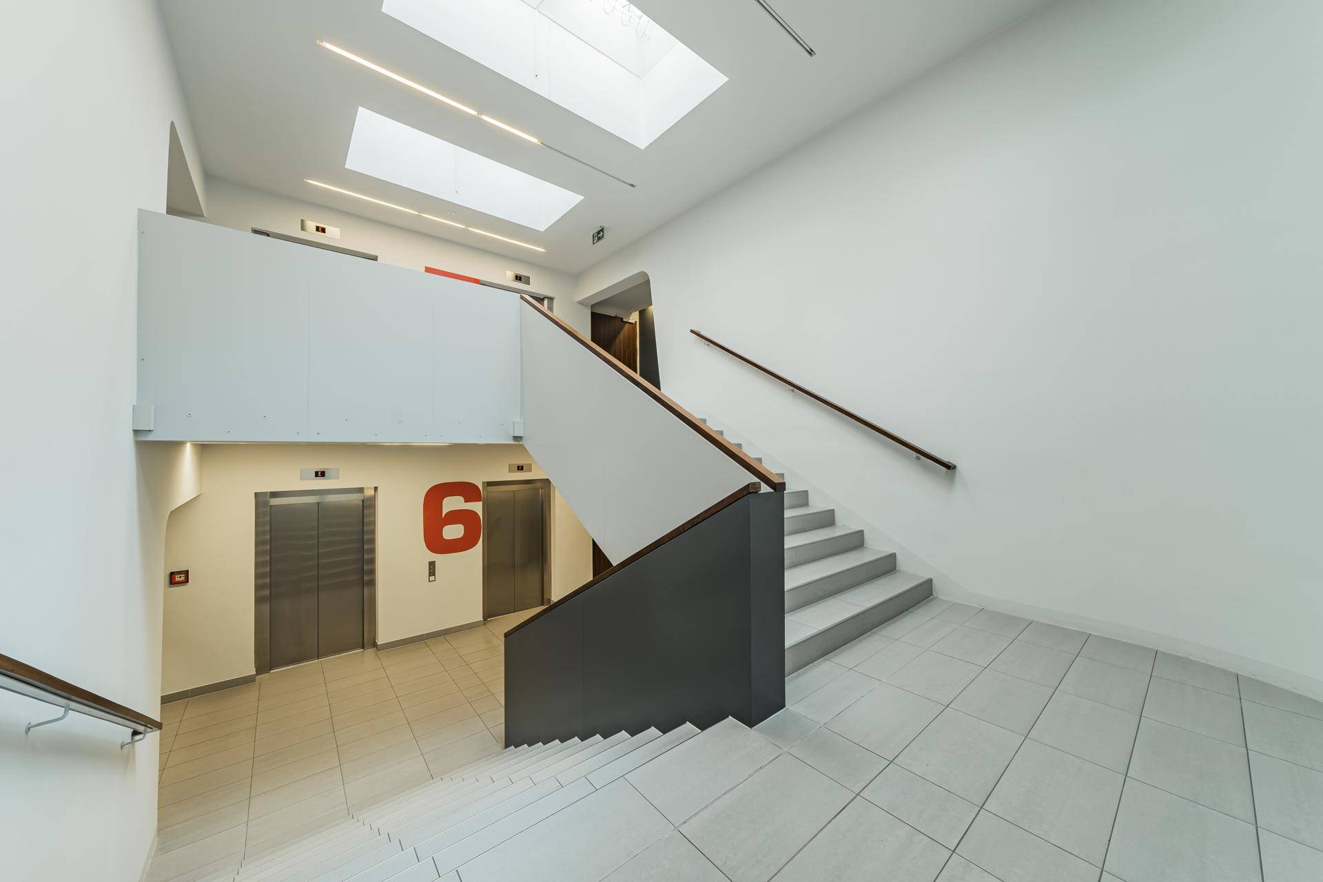 Moderner Treppenaufgang mit Fliesenboden, Handläufen und zwei Aufzügen.