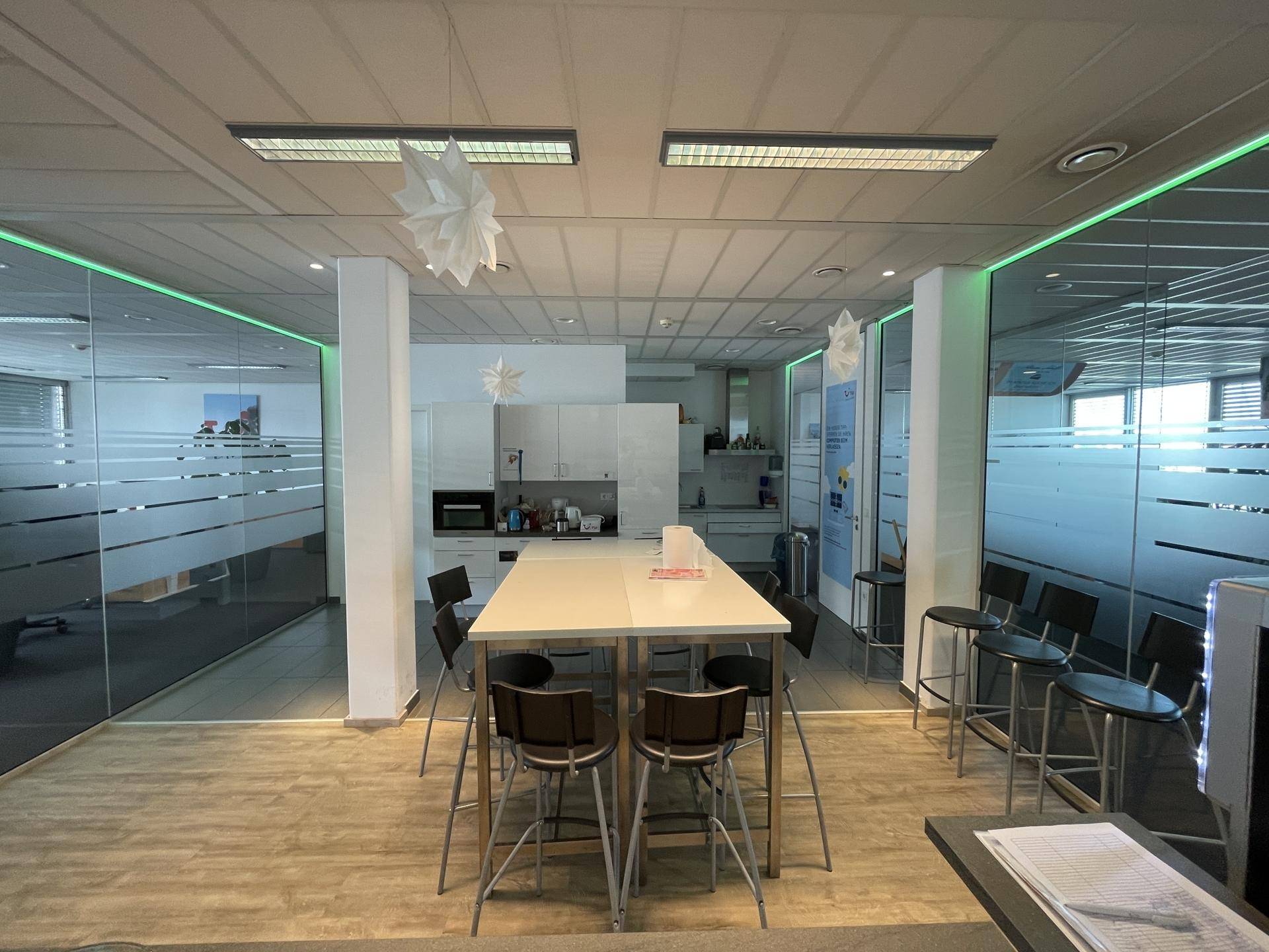 Moderne Büroküche und Essbereich mit langer Theke, Barhockern und Glasabtrennungen mit grüner Beleuchtung.