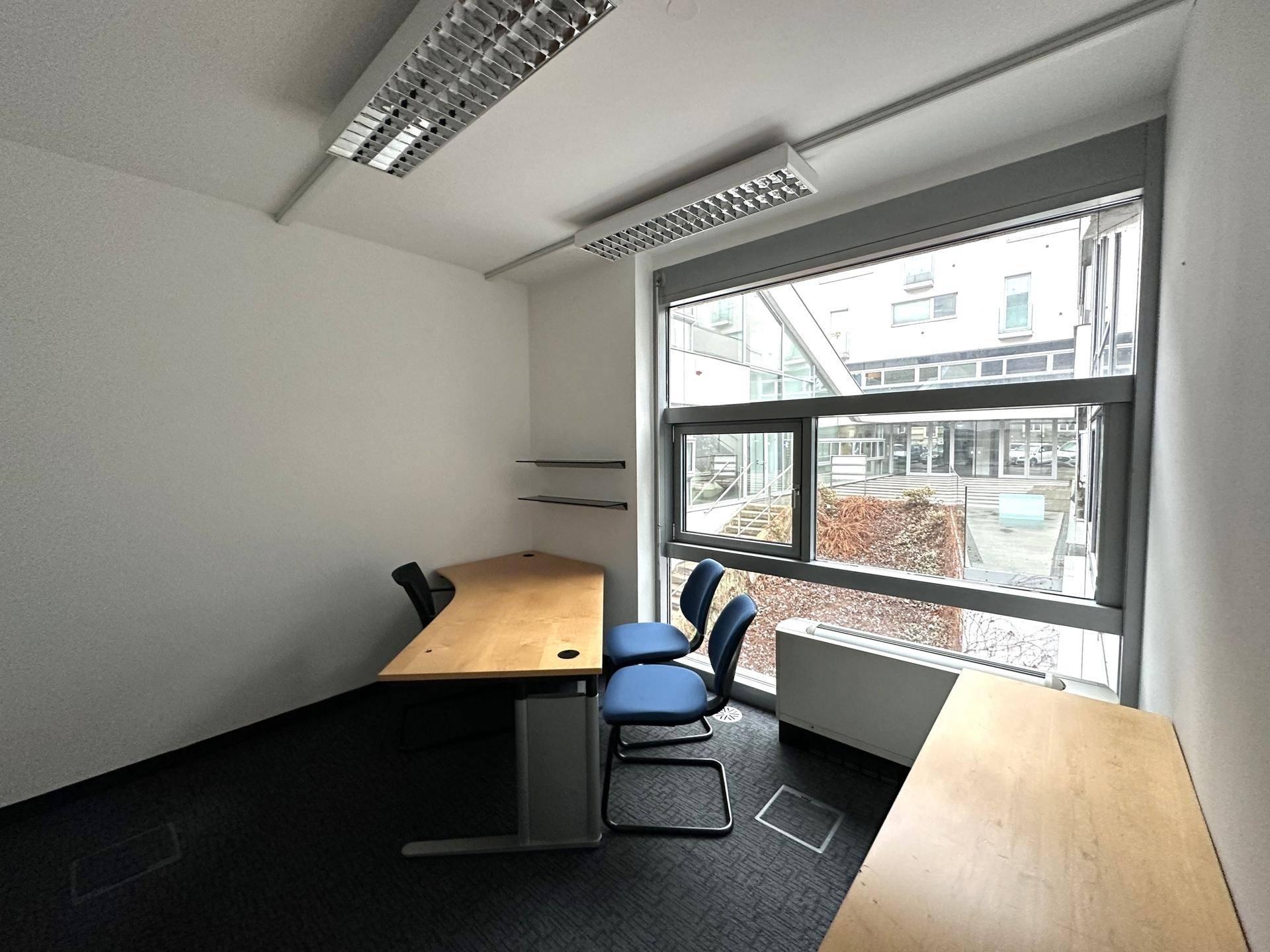 Helles Büro mit Teppichboden, großen Fenstern und einfacher Möblierung, bietet Blick auf den Innenhof.
