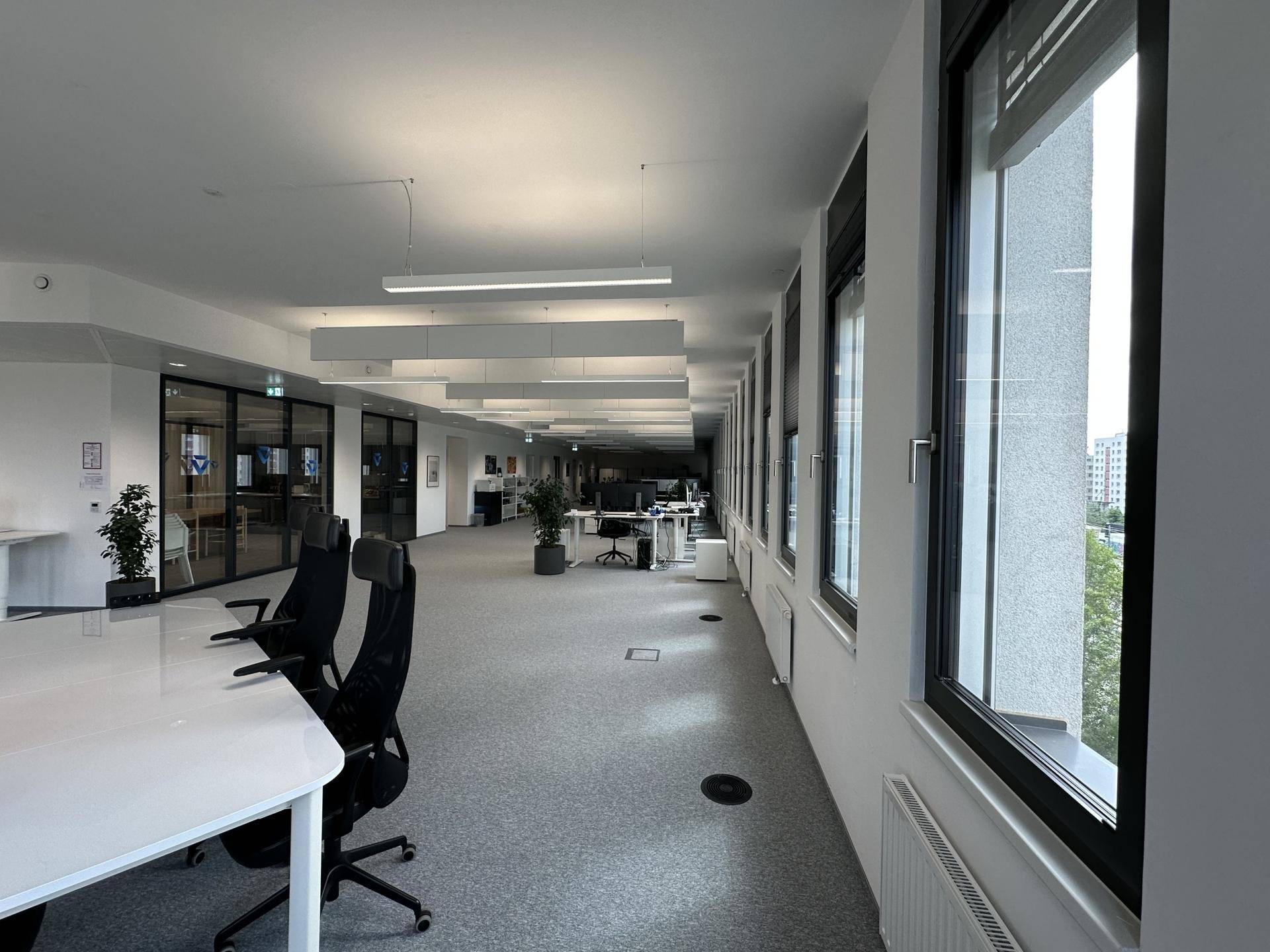 Langer, offener Bürobereich mit durchgehender Fensterfront, grauen Teppichböden und mehreren Arbeitsplätzen.