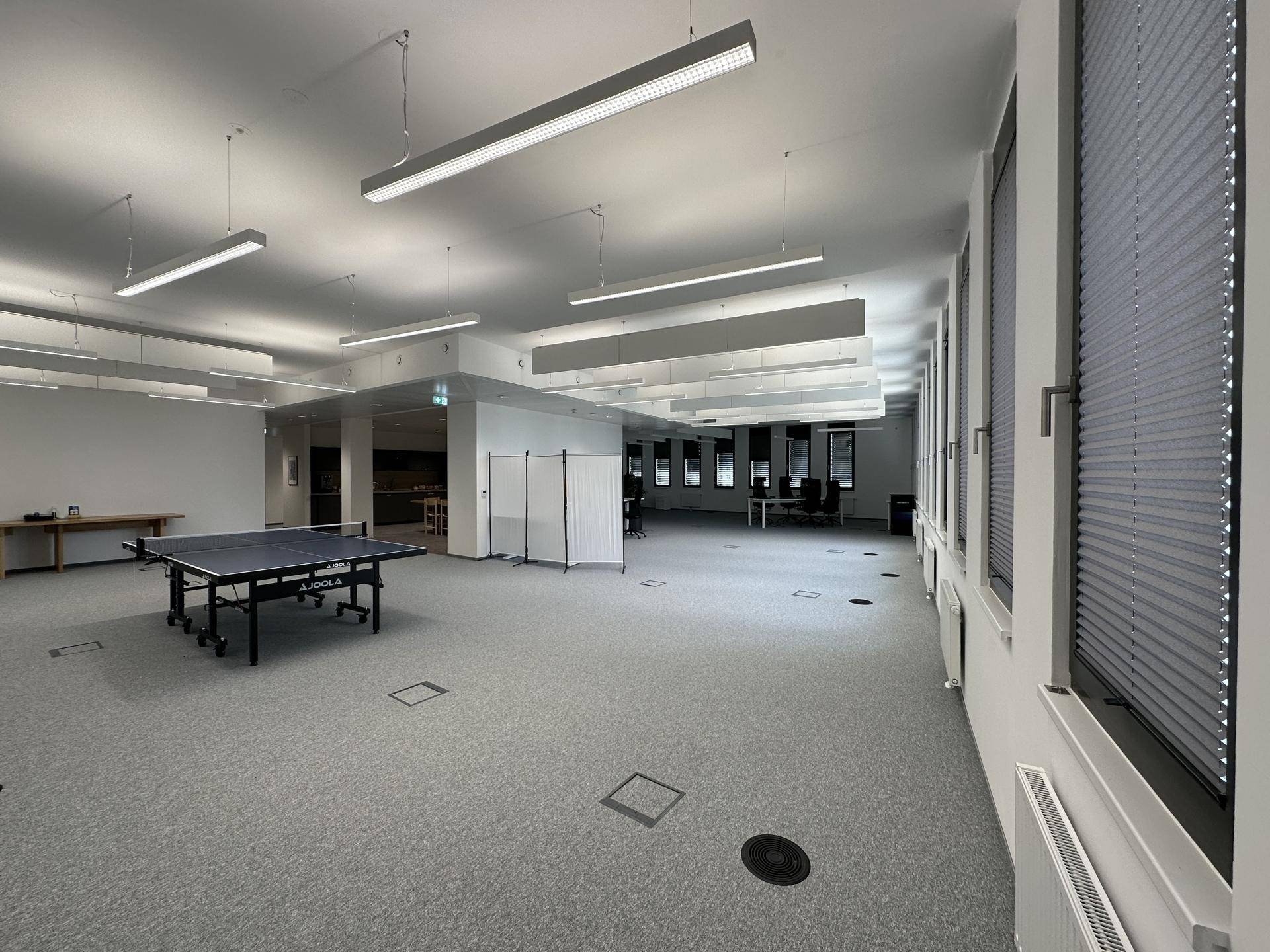 Großer offener Bürobereich mit grauen Teppichböden, vielen Fenstern und einem Tischtennistisch zur Entspannung.