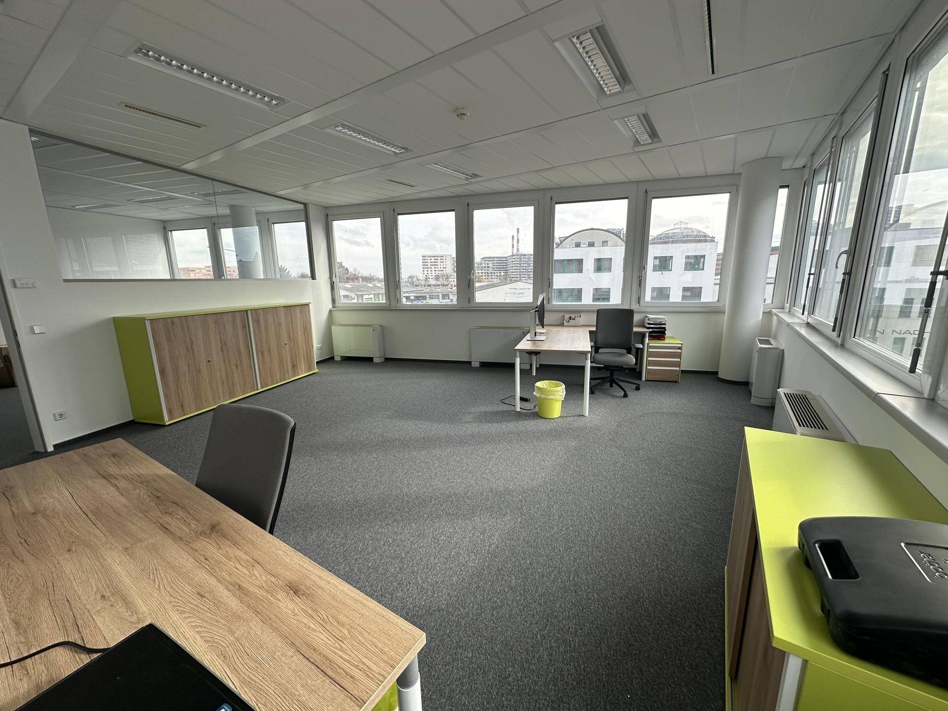 Geräumiges Büro mit Teppichboden, großen Fenstern und modernen Möbeln in Holz- und Grüntönen mit Stadtblick.