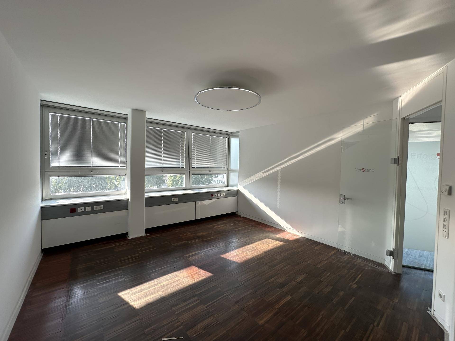 Büroraum mit dunklem Holzparkettboden, großen Fenstern mit Jalousien und einer modernen Glastür.