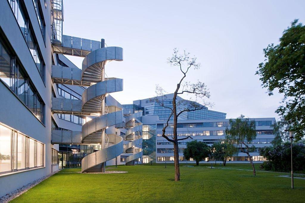 Gebäude mit markanter spiralförmiger Außentreppe, die zu einer gepflegten Rasenfläche führt, umgeben von moderner Architektur.