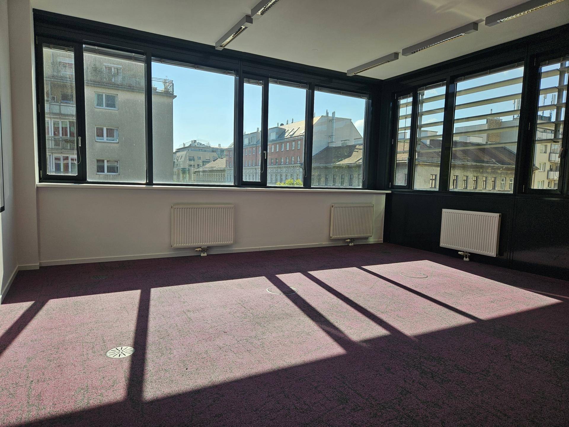 Leerer Büroraum mit großen Fenstern, die einen Blick auf die Stadt bieten, und lila Teppichboden.