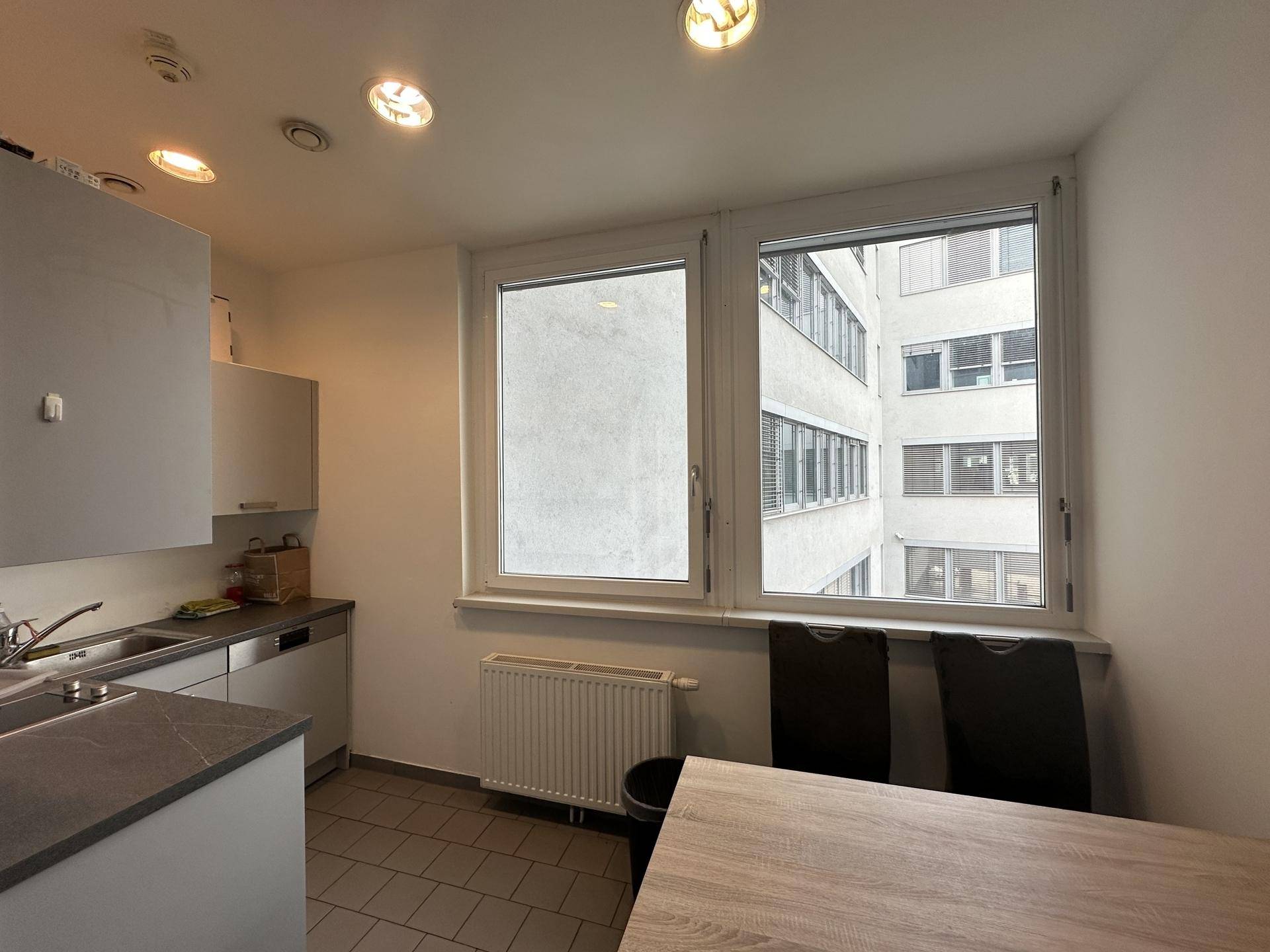 Kleine Küchenzeile mit Spüle, weißen Schränken und einem Esstisch, beleuchtet durch ein Fenster mit Hofblick.