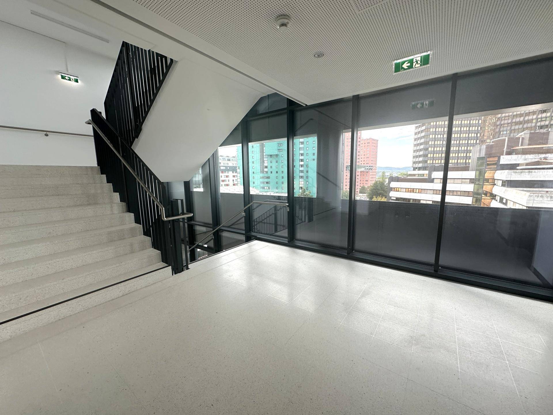 Modernes Treppenhaus mit großen Fenstern, schwarzem Geländer und Blick auf die Stadt.