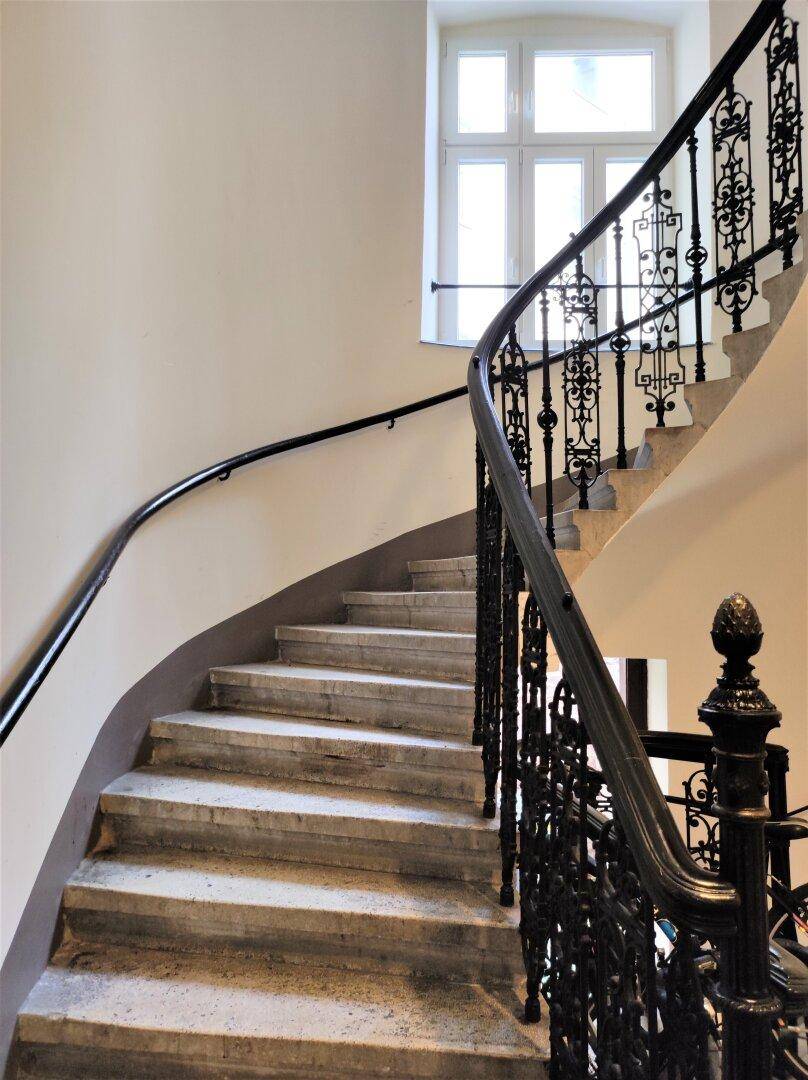 Elegantes Treppenhaus mit geschwungener Steintreppe und kunstvollem schmiedeeisernem Geländer, hell.