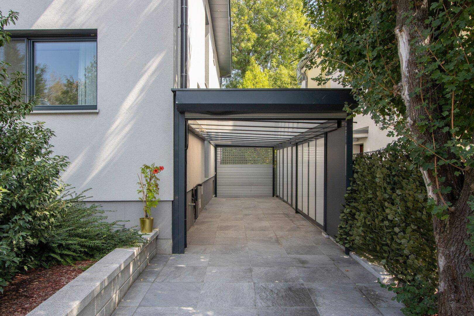 Langer überdachter Carport mit Fliesenboden und Zugang zum Haus, umgeben von Grün.
