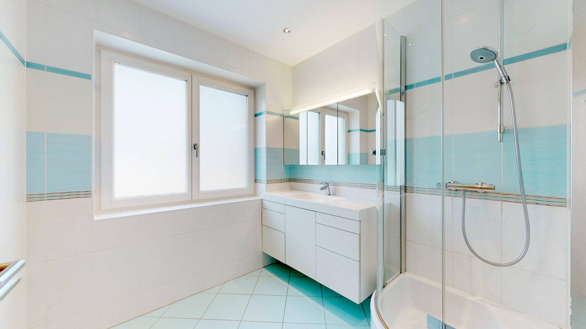 Helles Badezimmer mit Dusche, Einzelwaschbecken und Fenster, in weiß-blauer Farbgebung.