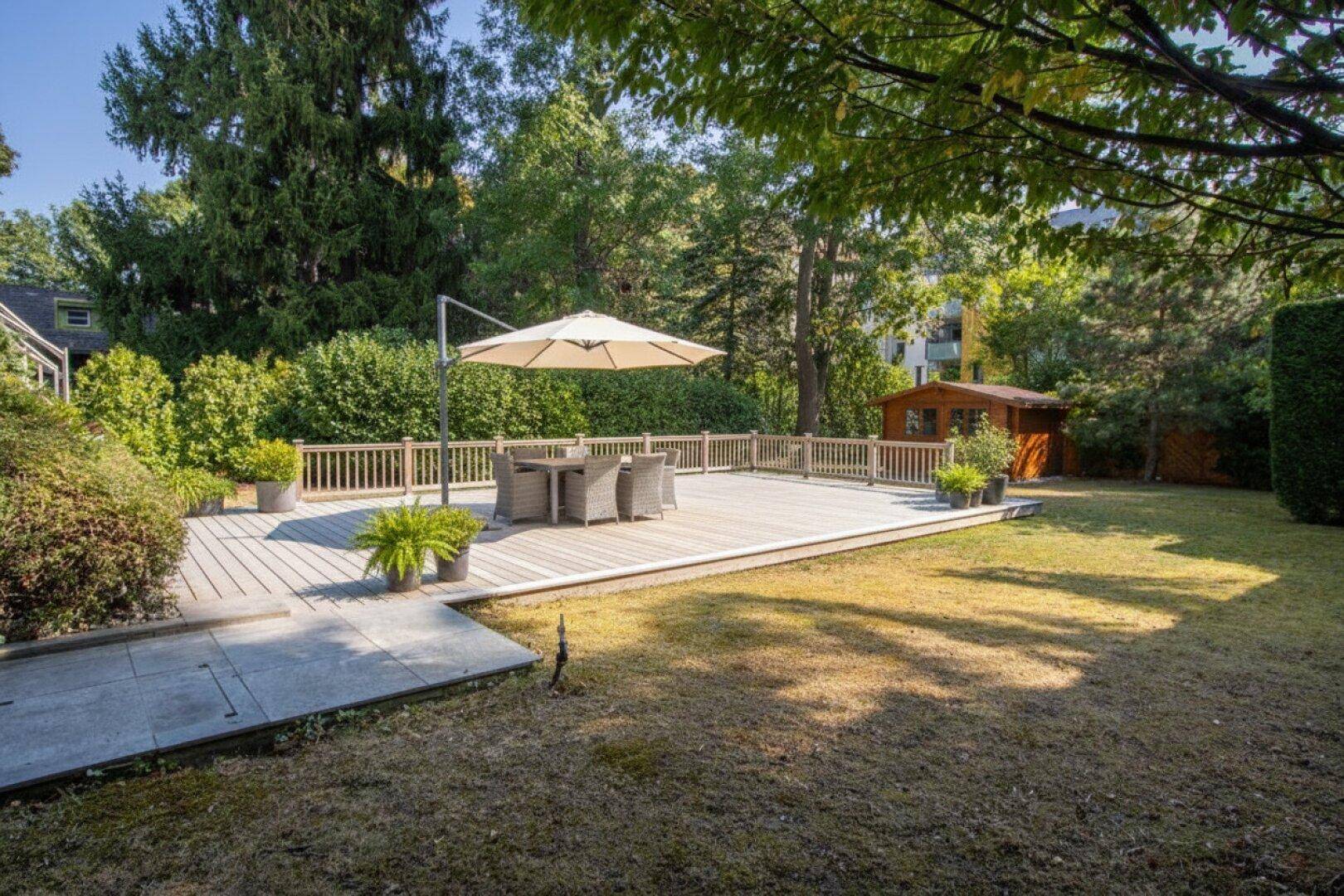 Große Holzterrasse im Garten mit Esstisch und Sonnenschirm, umgeben von viel Grün.