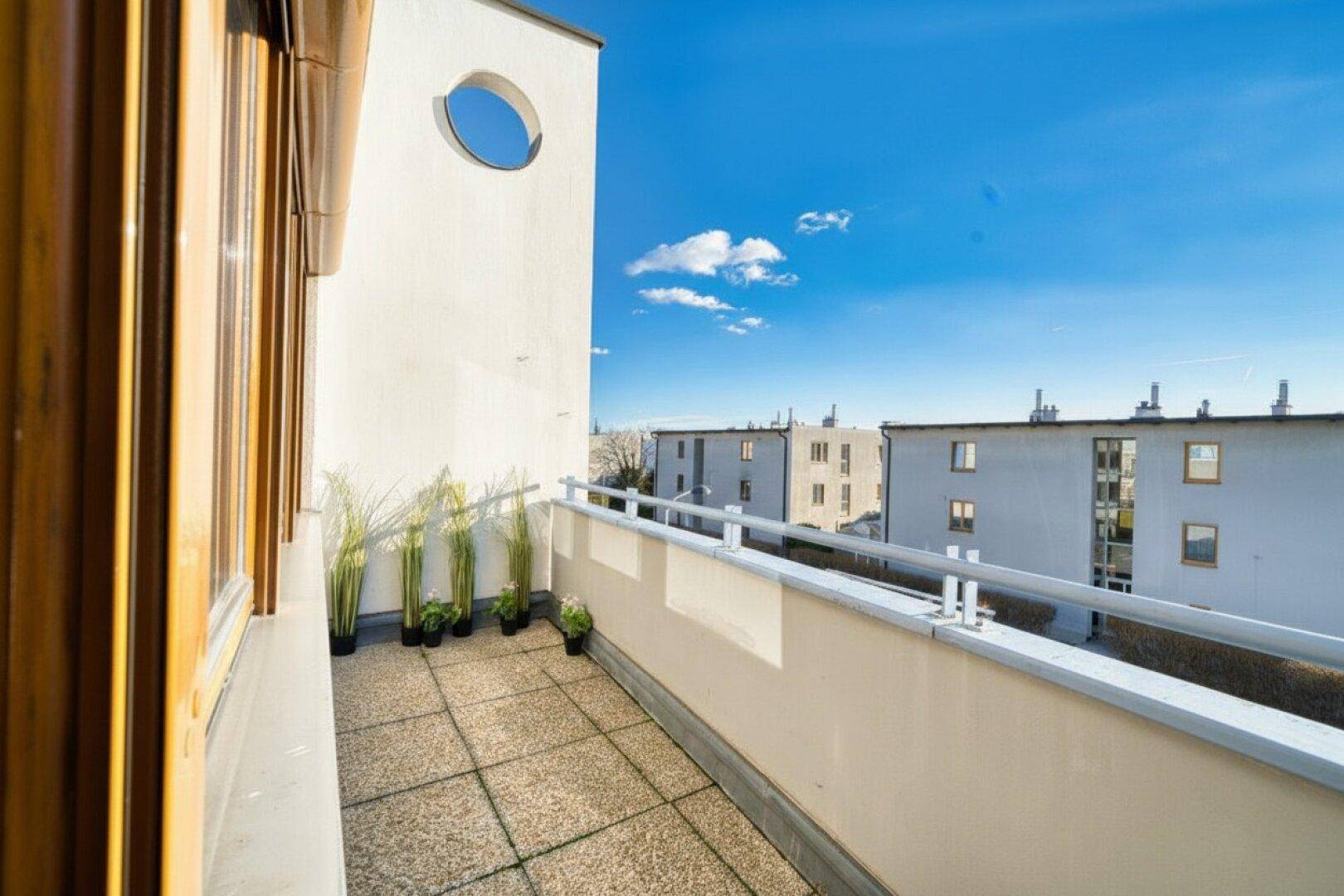 Unmöblierter Balkon mit Fliesenboden und Blick auf umliegende Gebäude unter blauem Himmel.