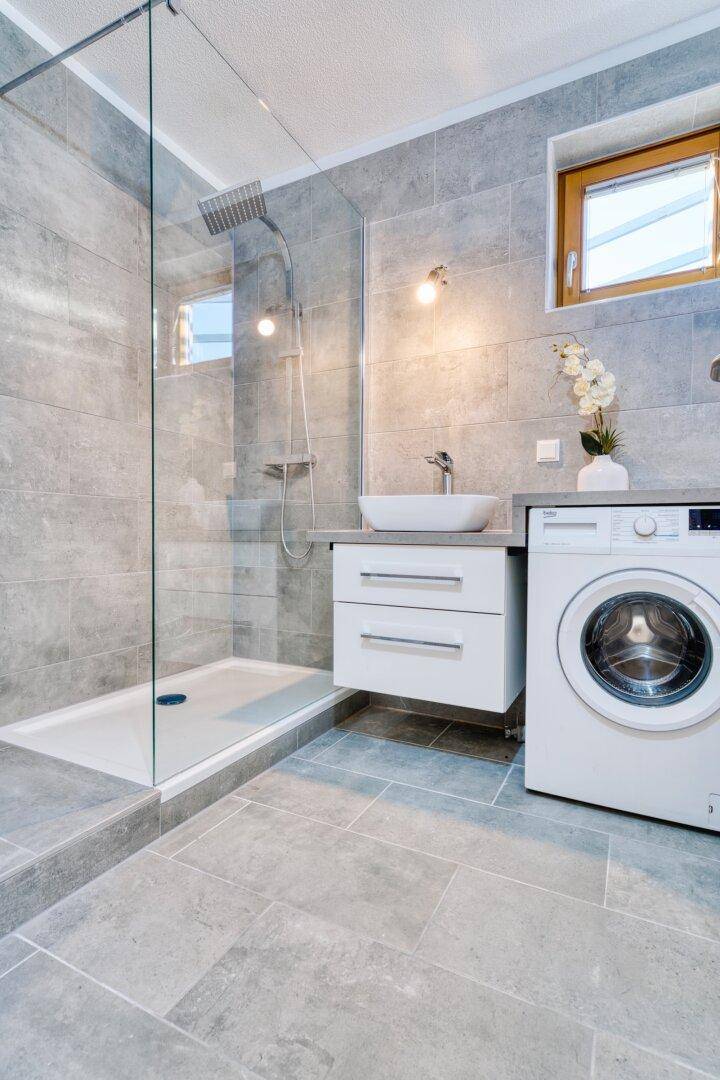 Modernes Badezimmer mit grauen Fliesen, ebenerdiger Dusche und Waschmaschine.