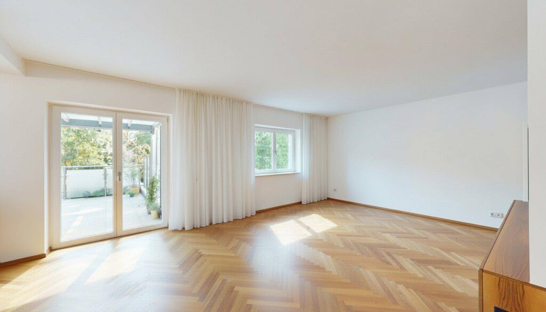 Geräumiges, leeres Wohnzimmer mit Fischgrätparkett und großen Fenstern, die viel Licht hereinlassen.