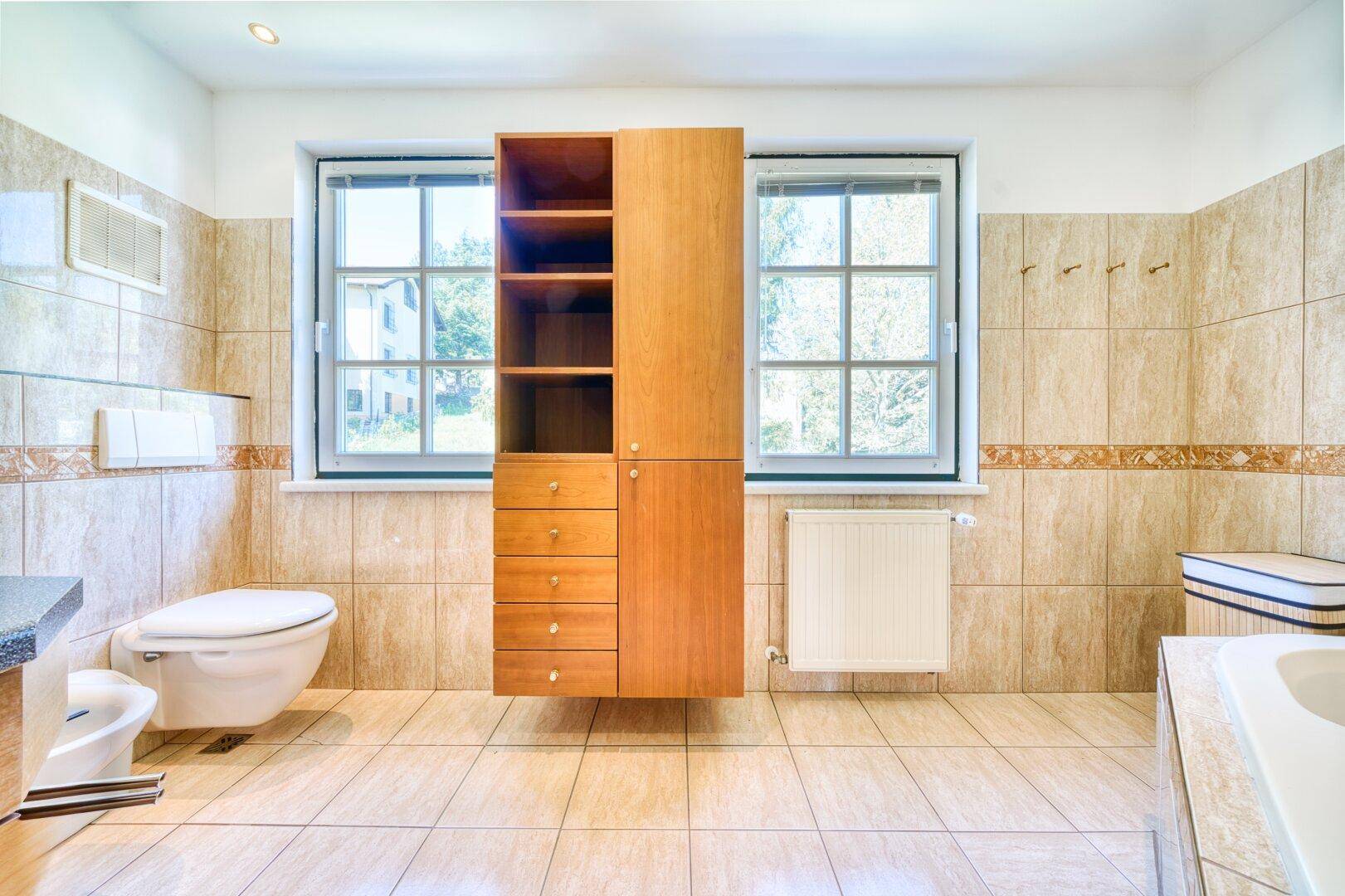Badezimmer mit beigefarbenen Fliesen, wandhängendem WC und zwei Fenstern.