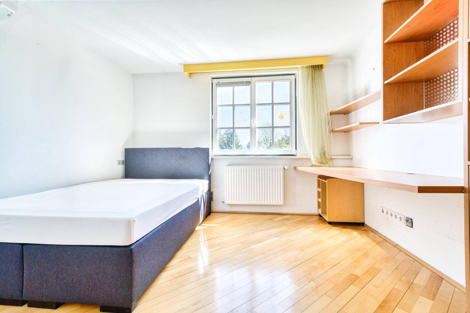 Helles Schlafzimmer mit Parkettboden, Fenster und integriertem Schreibtischbereich.