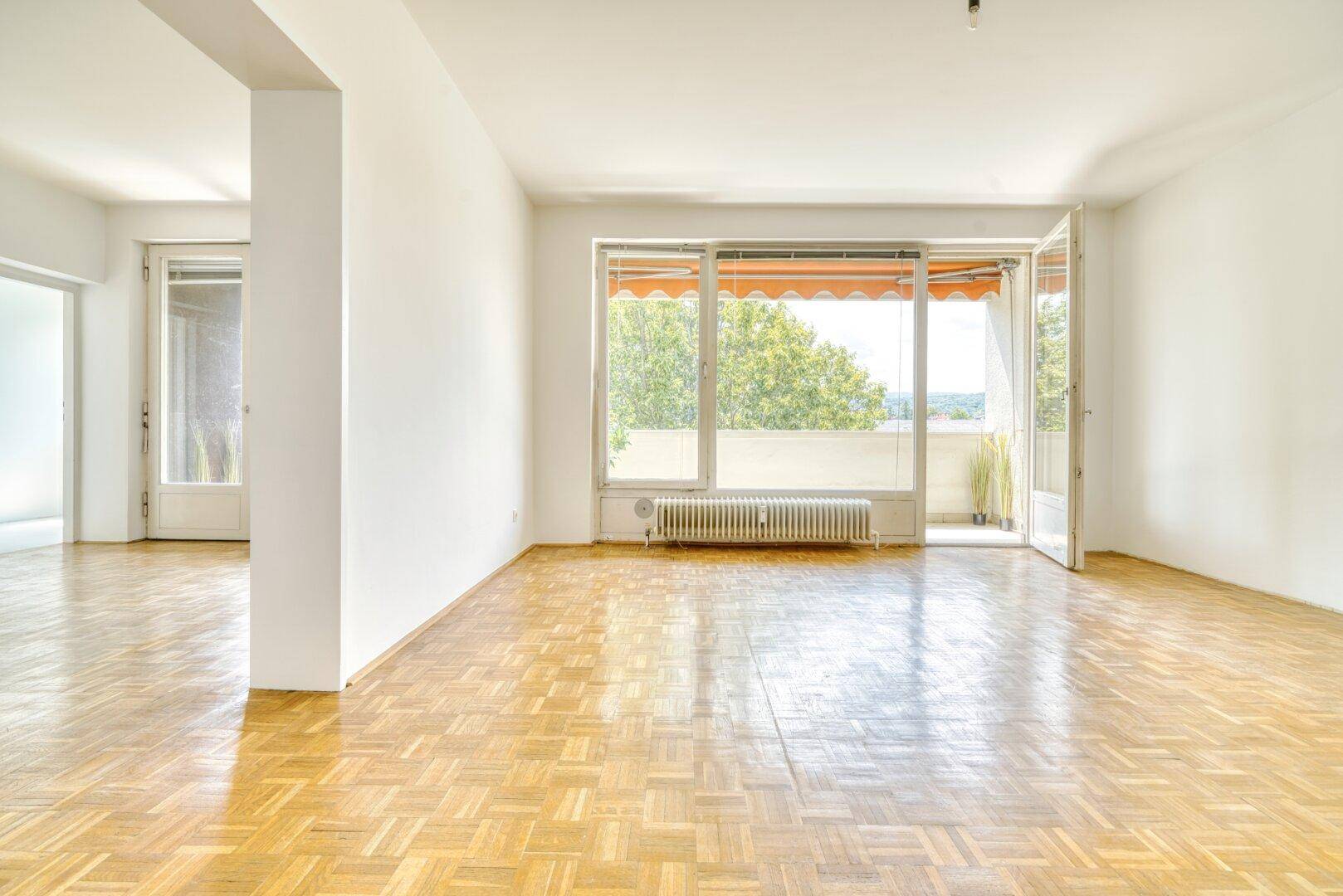 Großzügiges Wohnzimmer mit Parkettboden und breiter Fensterfront zum Balkon.