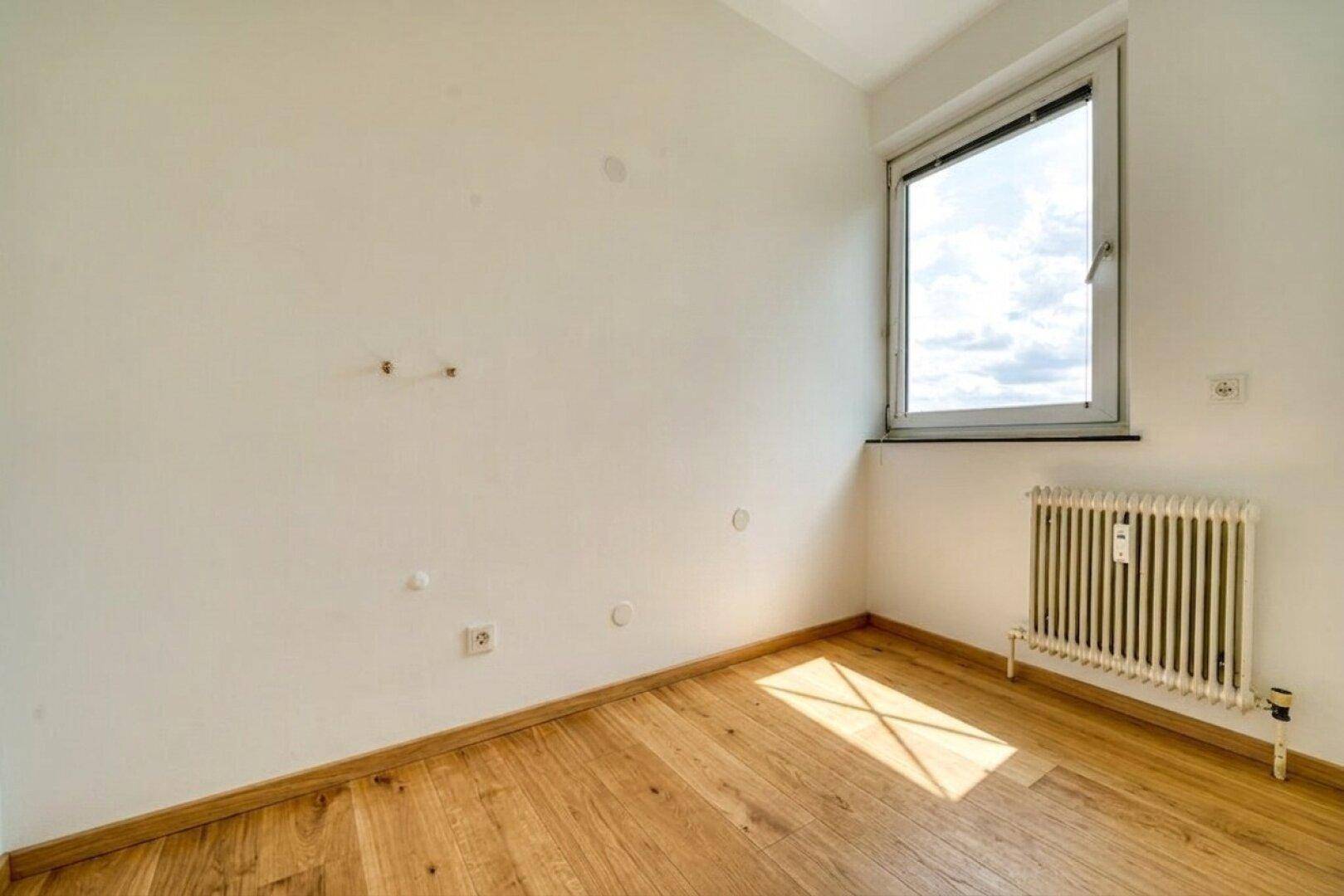 Leeres Zimmer mit hellem Holzfußboden, Fenster und Heizkörper, durchflutet von natürlichem Licht.