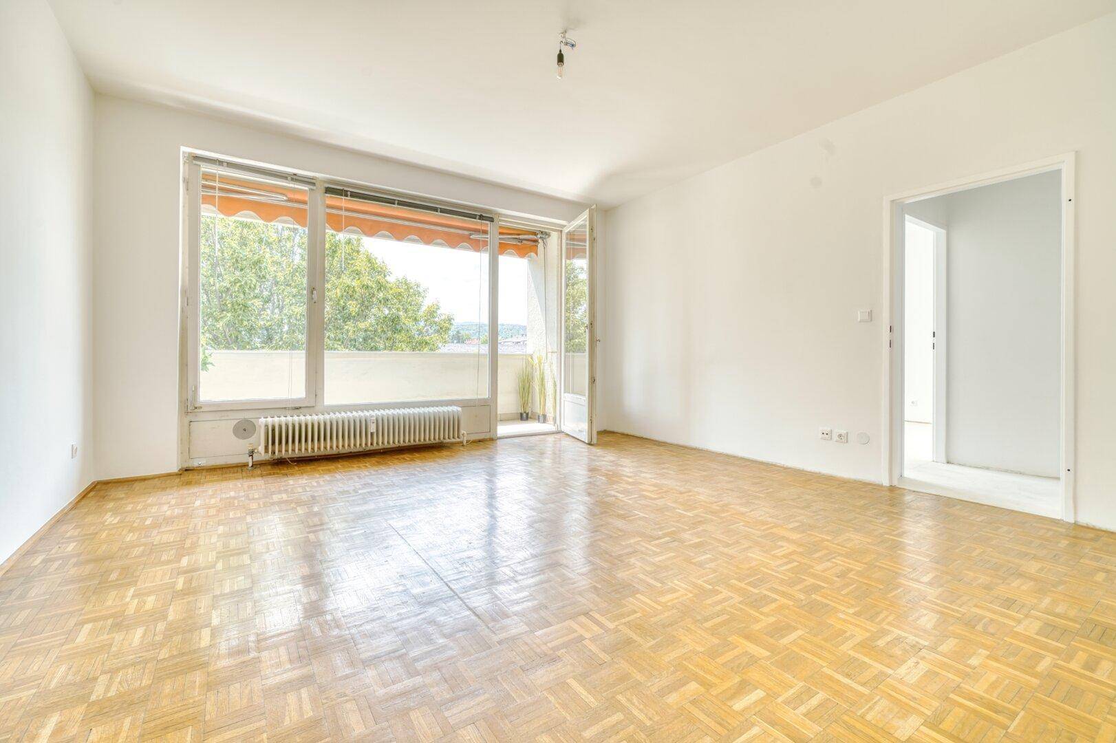 Leeres Wohnzimmer mit Parkettboden und breiter Fensterfront zum Balkon.