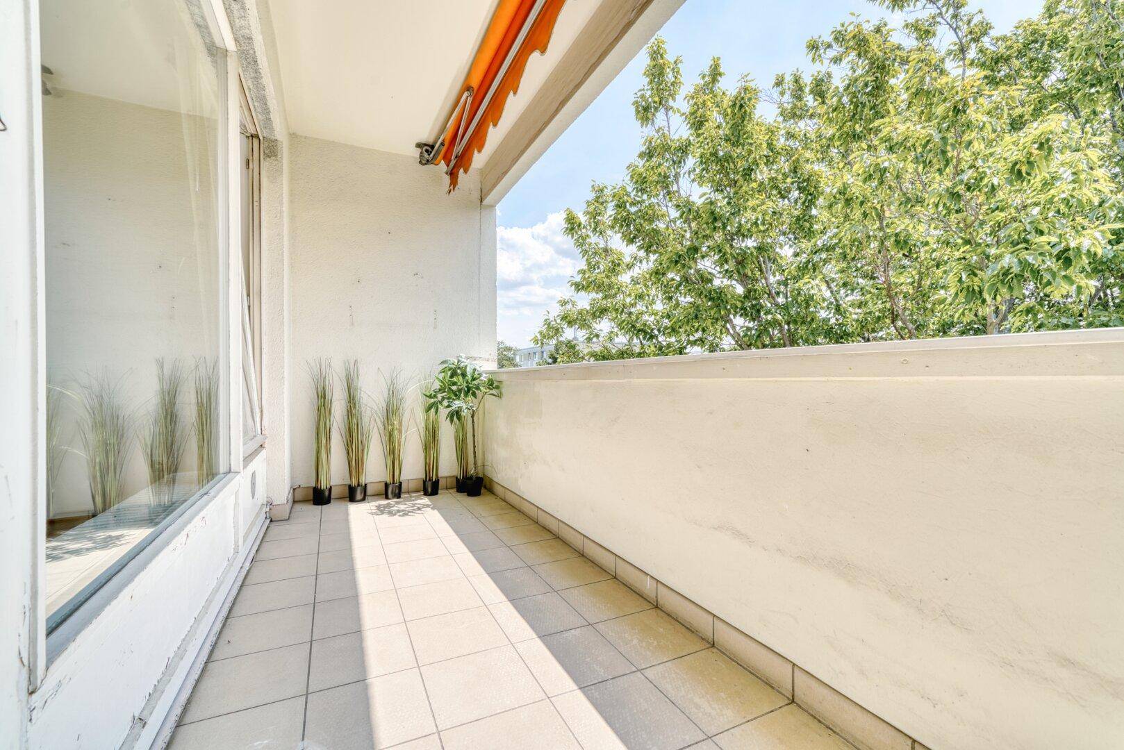 Heller Balkon mit Fliesenboden und Markise, umgeben von grünen Bäumen.